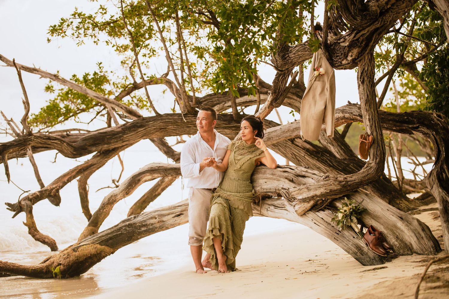 Couple posing together on large driftwood tree at sandy beach.