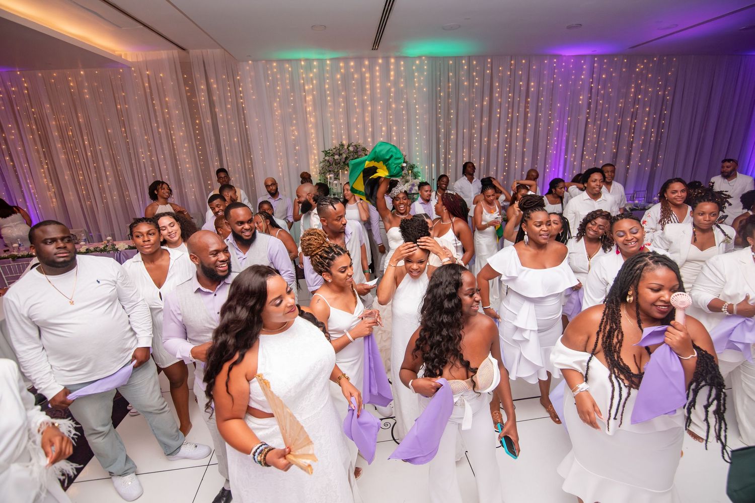 Wedding celebration with guests dressed in all-white outfits dancing together under decorative lighting.