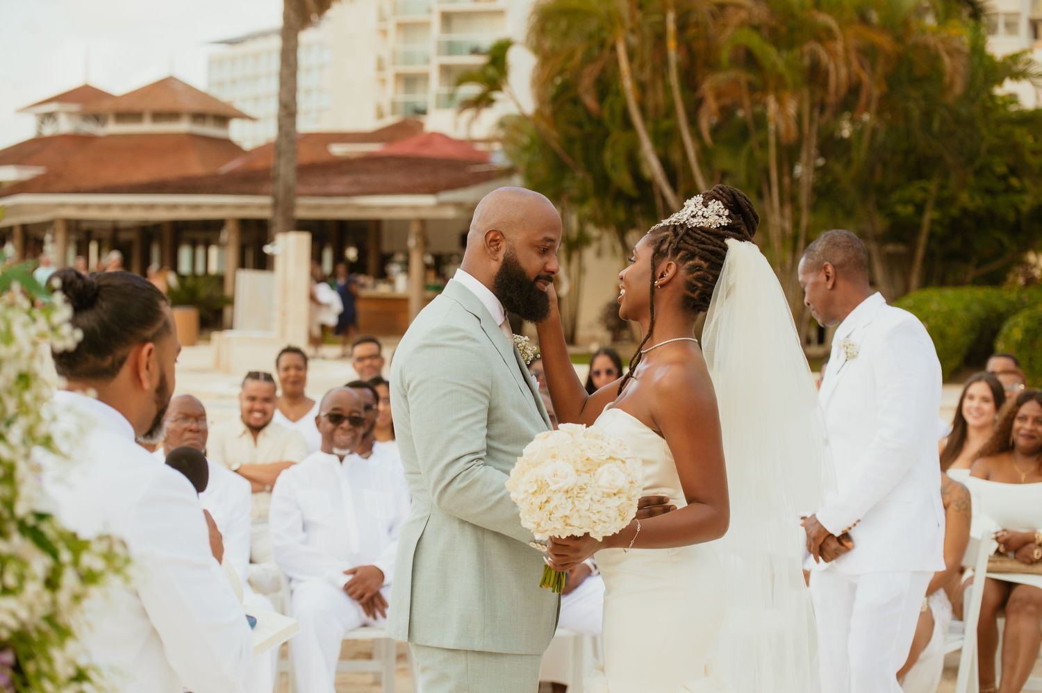 Beach wedding ceremony with tropical landscaping and resort buildings in the background.