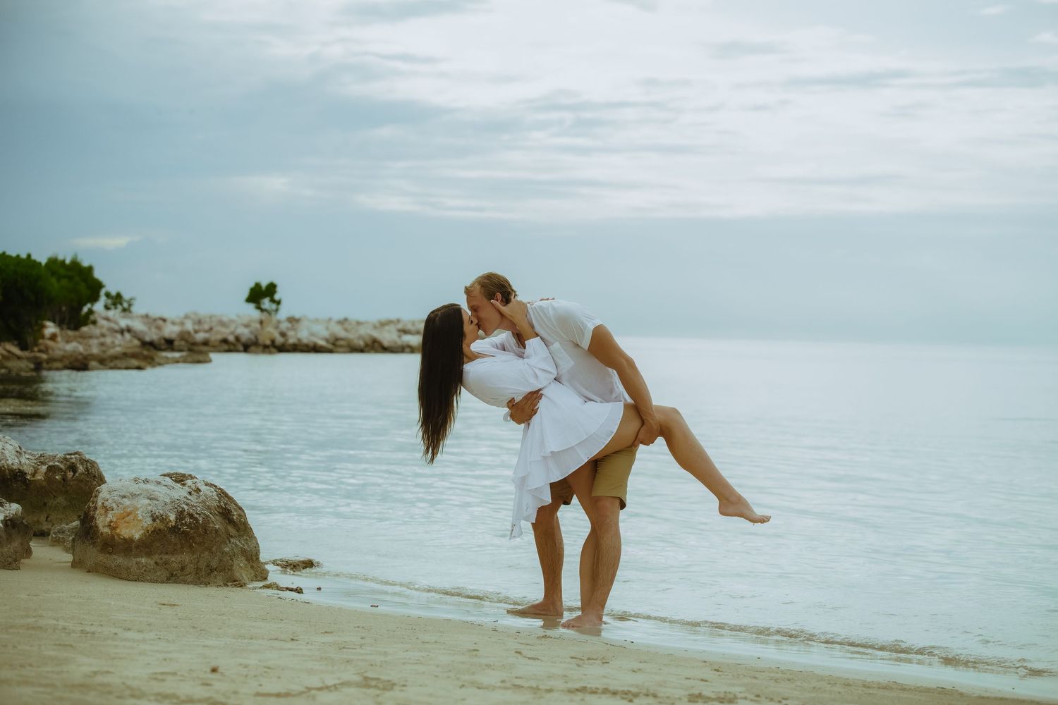 A playful couple in white clothing embraces on a scenic beach with rocks and ocean views.