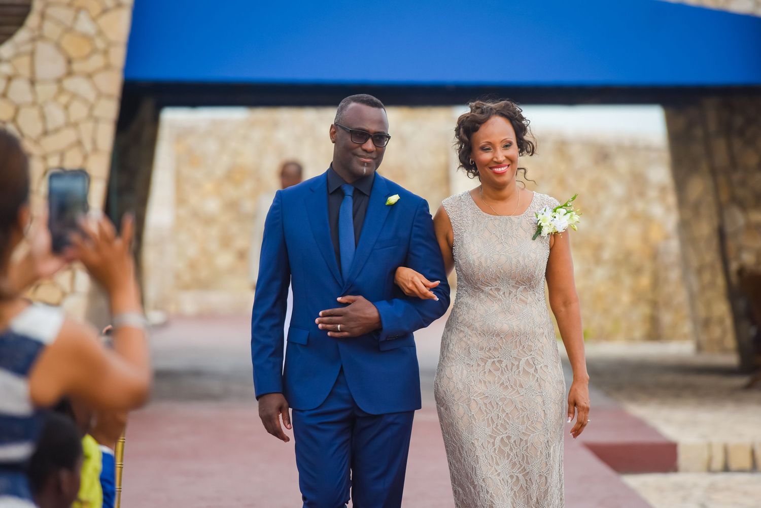 Wedding couple walking down outdoor aisle with stone wall backdrop.