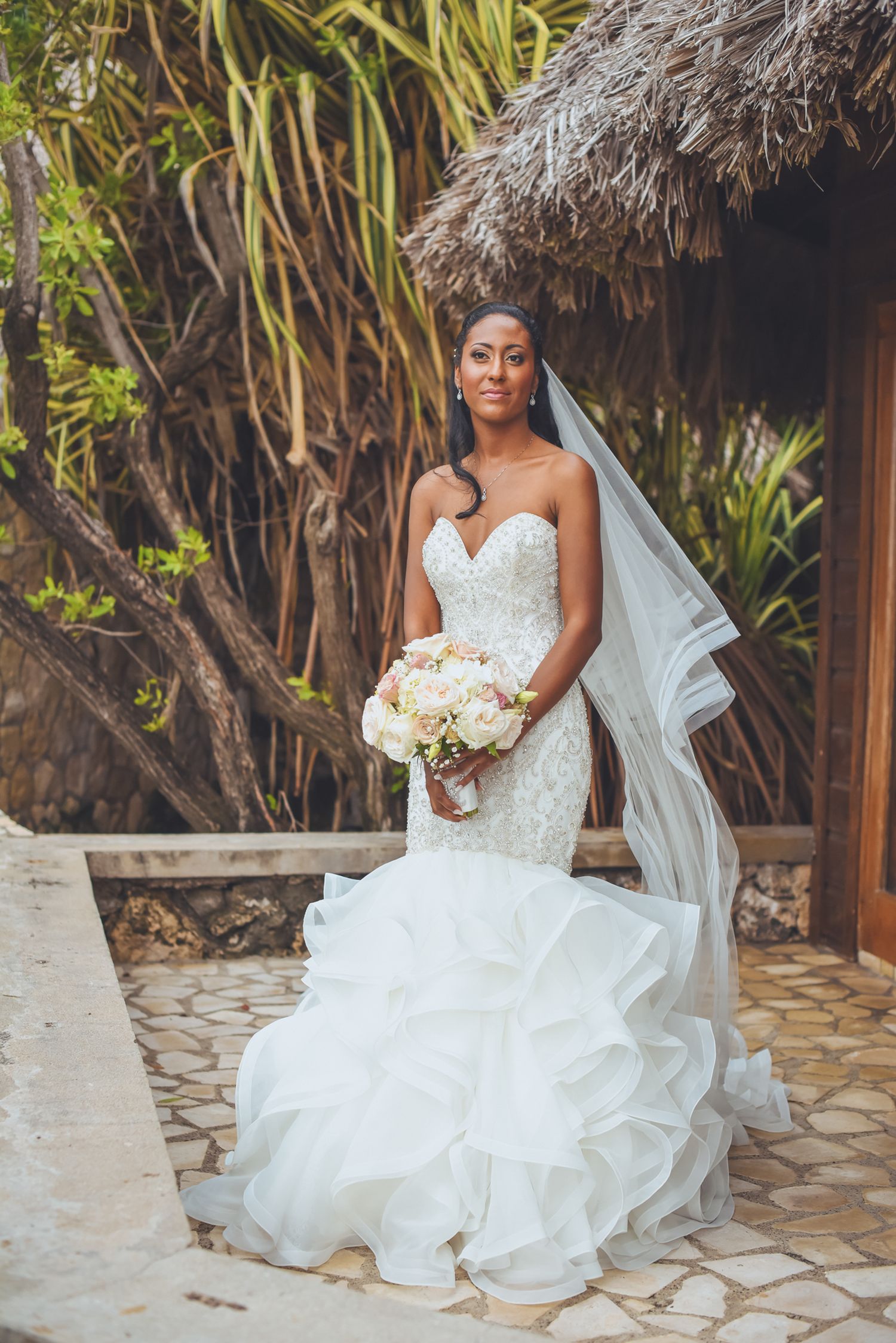 Bride in an embellished white gown poses under a wooden pergola with lush greenery.