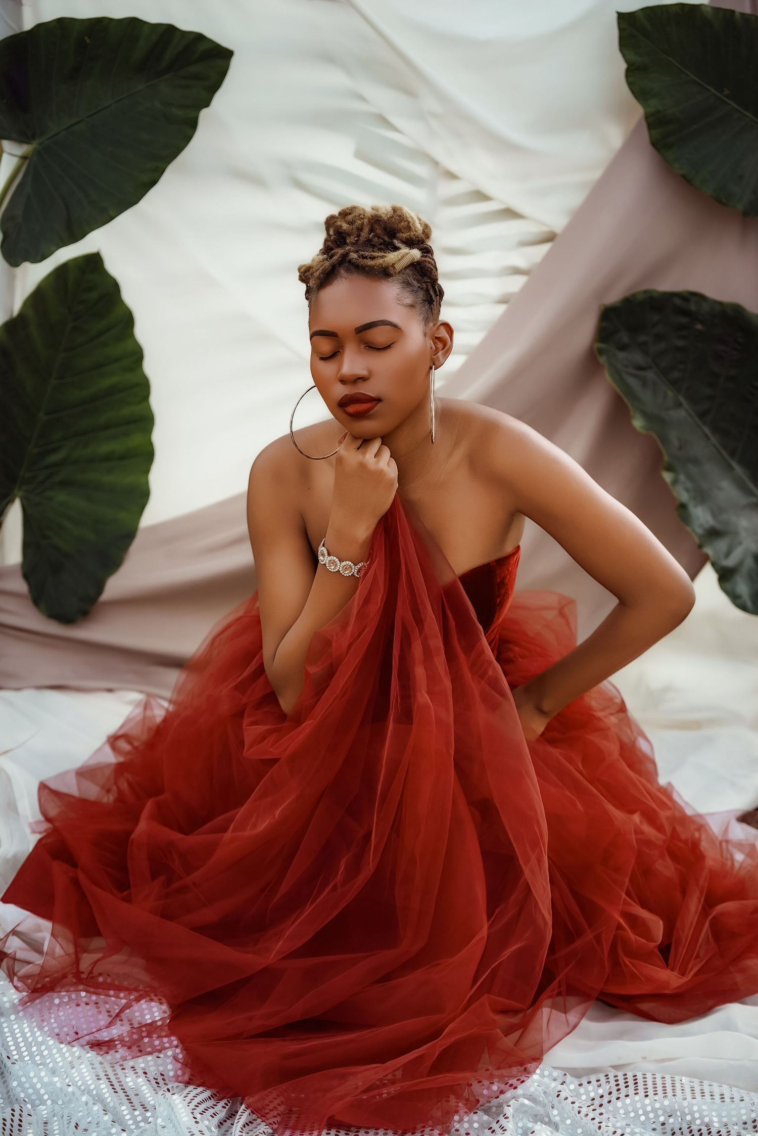 Elegant portrait featuring flowing red tulle gown against white backdrop with tropical leaves.