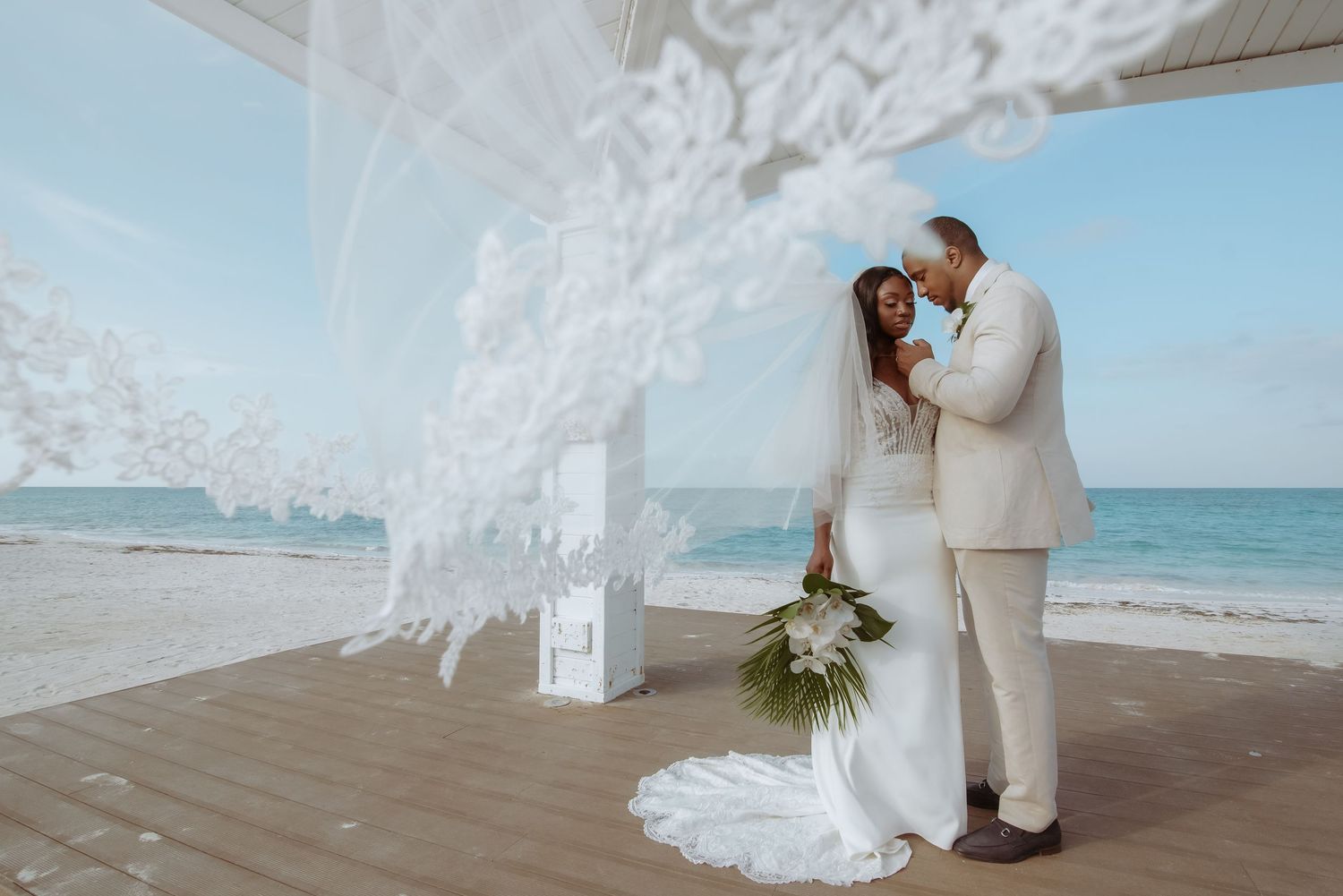 A couple shares a romantic moment under white lace decorations on a beachfront wooden deck during their wedding ceremony.