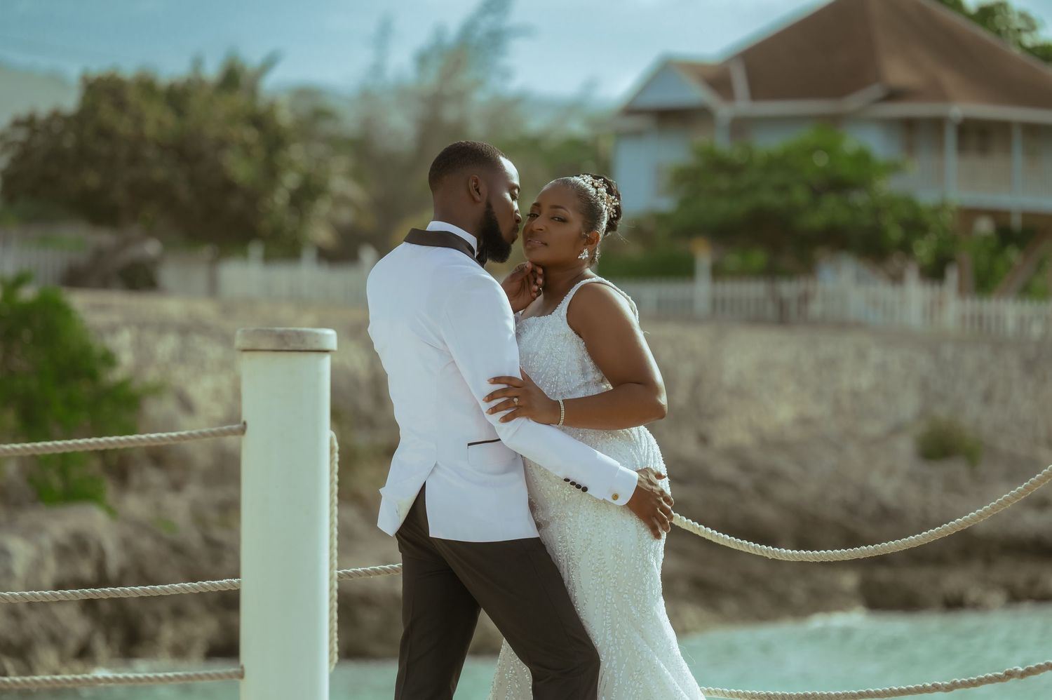 A romantic couple in white formal attire embrace on a beachfront deck during a tropical wedding celebration.