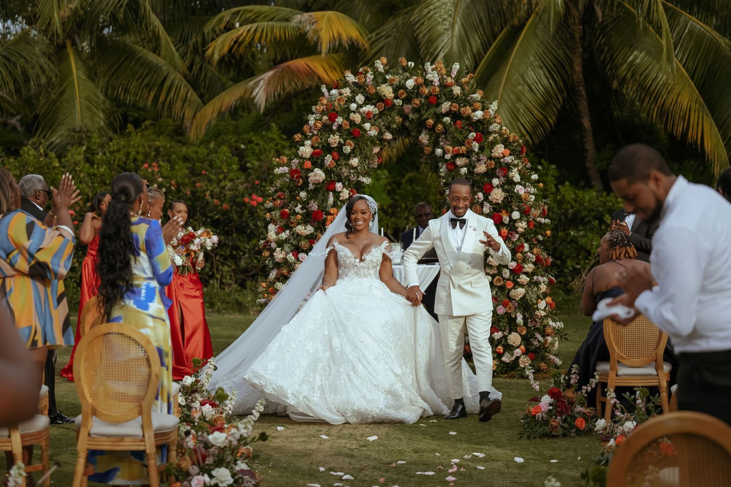 Wedding ceremony in progress at a lush tropical garden setting with floral arch and seated guests.
