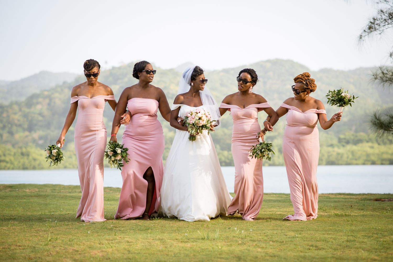 Bridesmaids in pink dresses pose together on a scenic hilltop overlooking a lake.