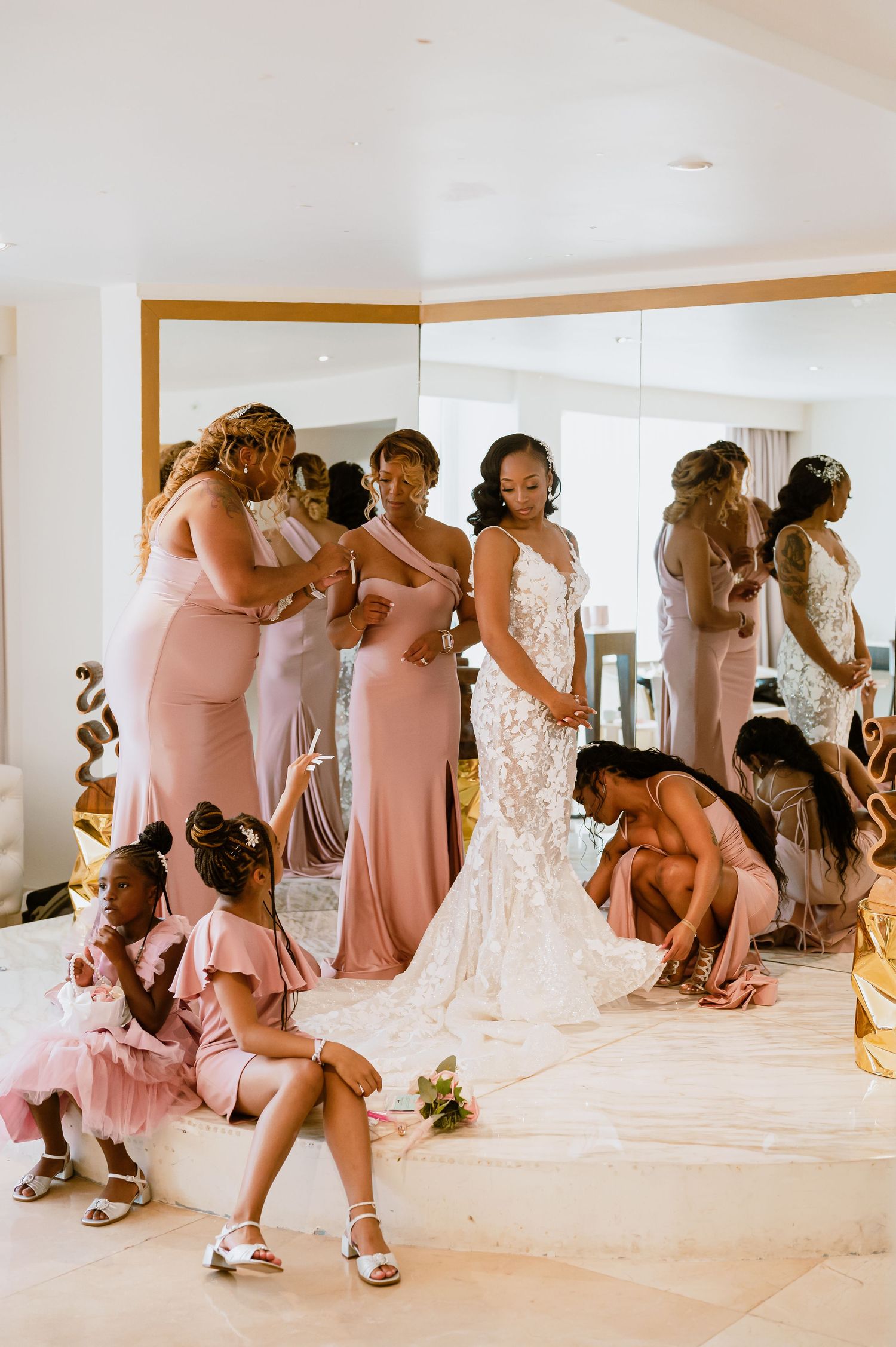 Bridesmaids in pink dresses help with wedding preparations in a sunlit room.