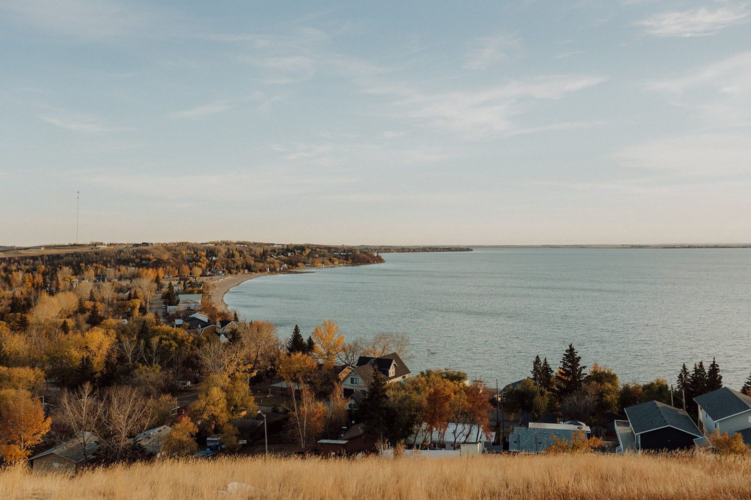 Aerial view of autumn trees and houses along a scenic lake or river shoreline at sunset.