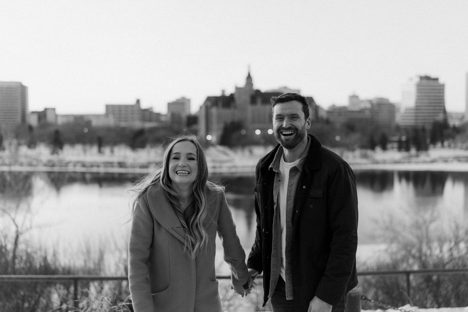 Couple standing by a lake with city skyline in the background during winter.