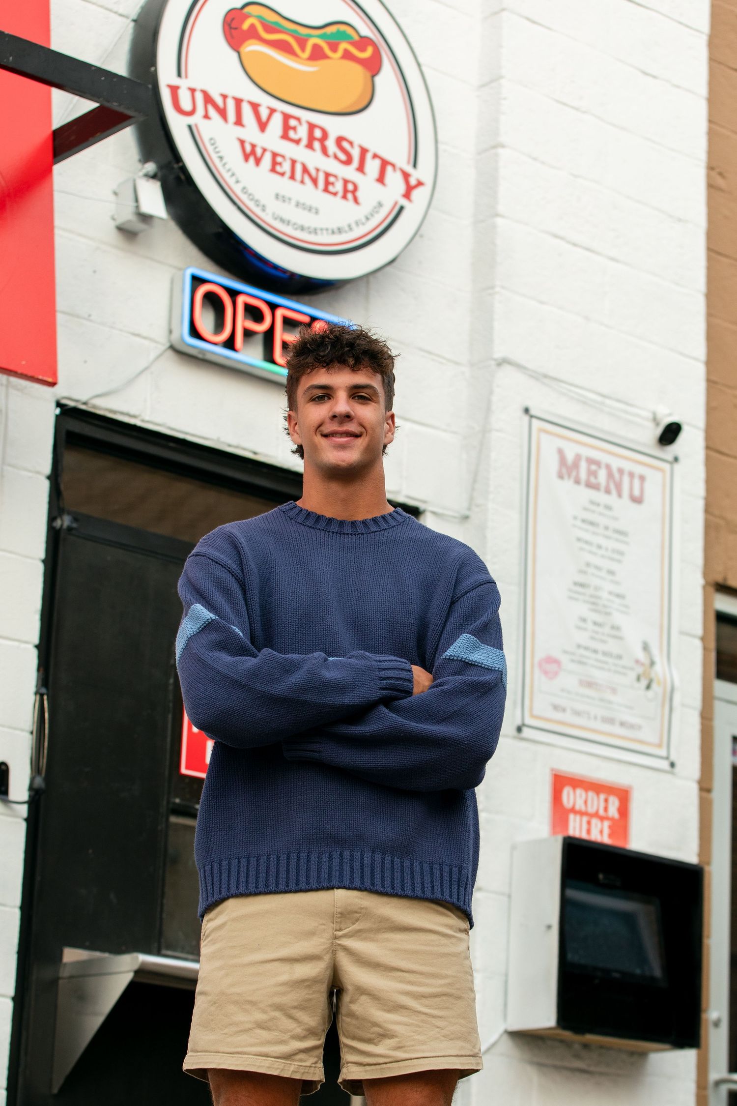 Students stand outside a University Weiner restaurant storefront wearing casual attire.