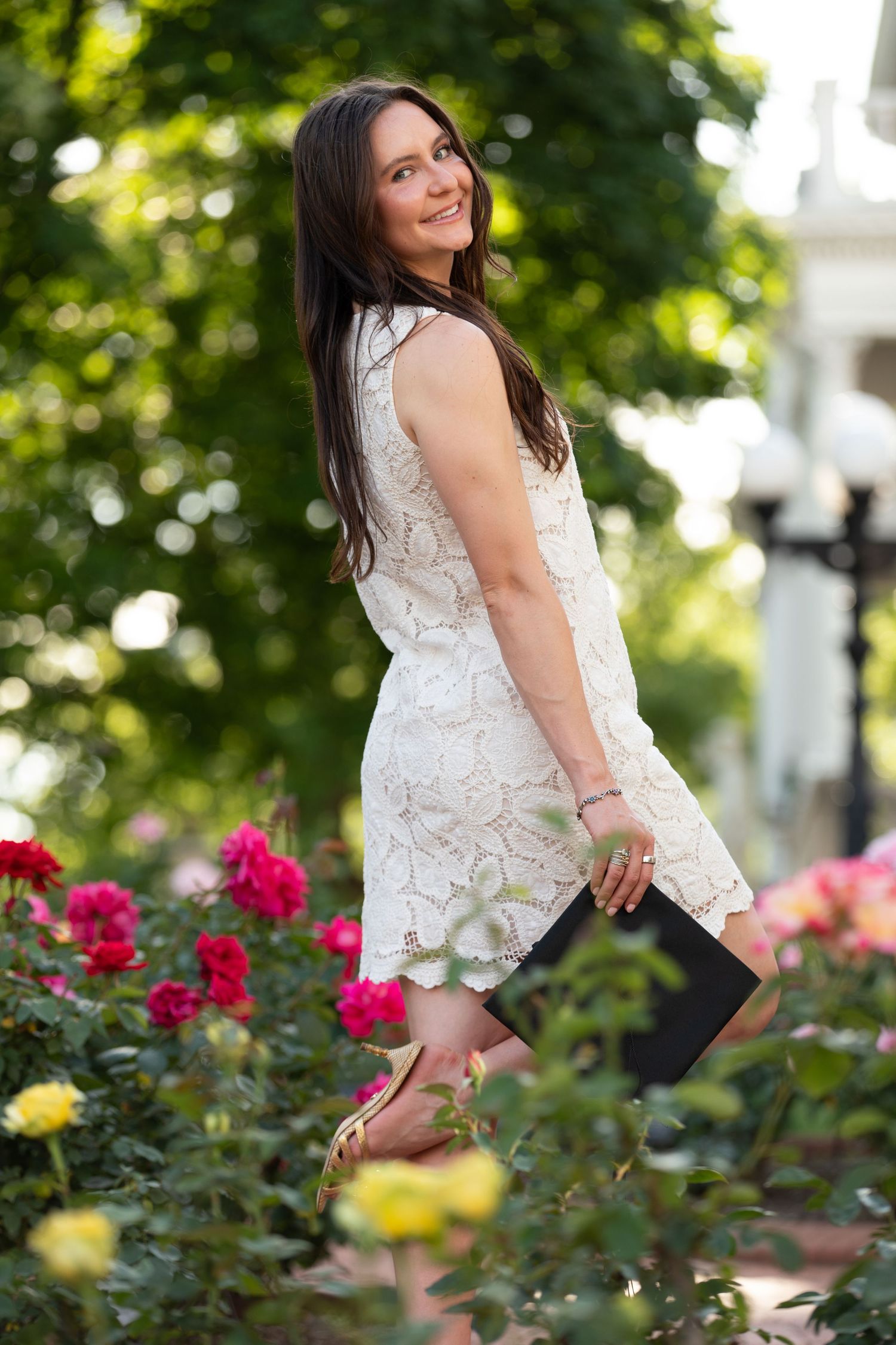 Woman in white lace dress poses in garden with blooming roses.