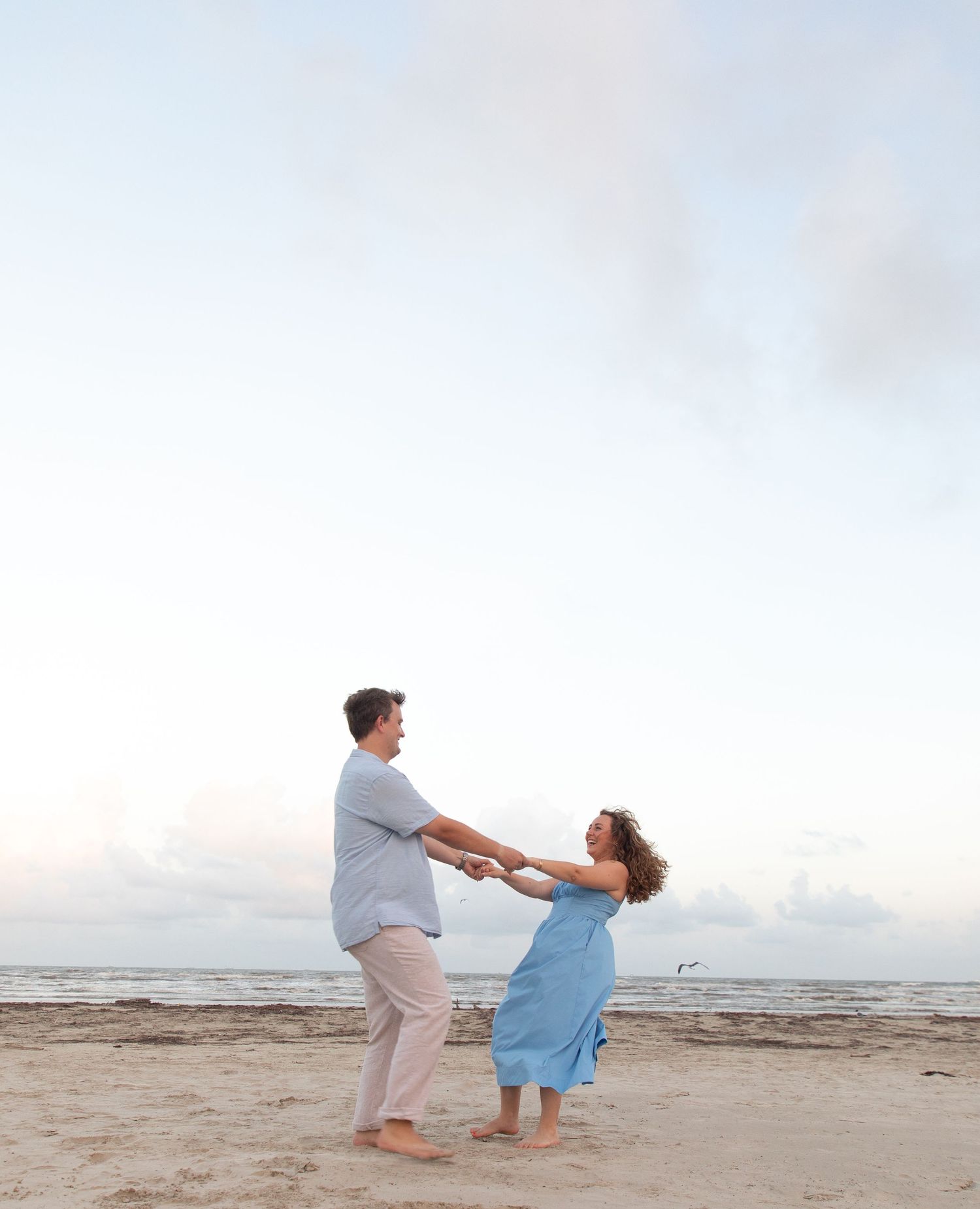 Couple playfully dancing and twirling together on sandy beach with ocean in background.