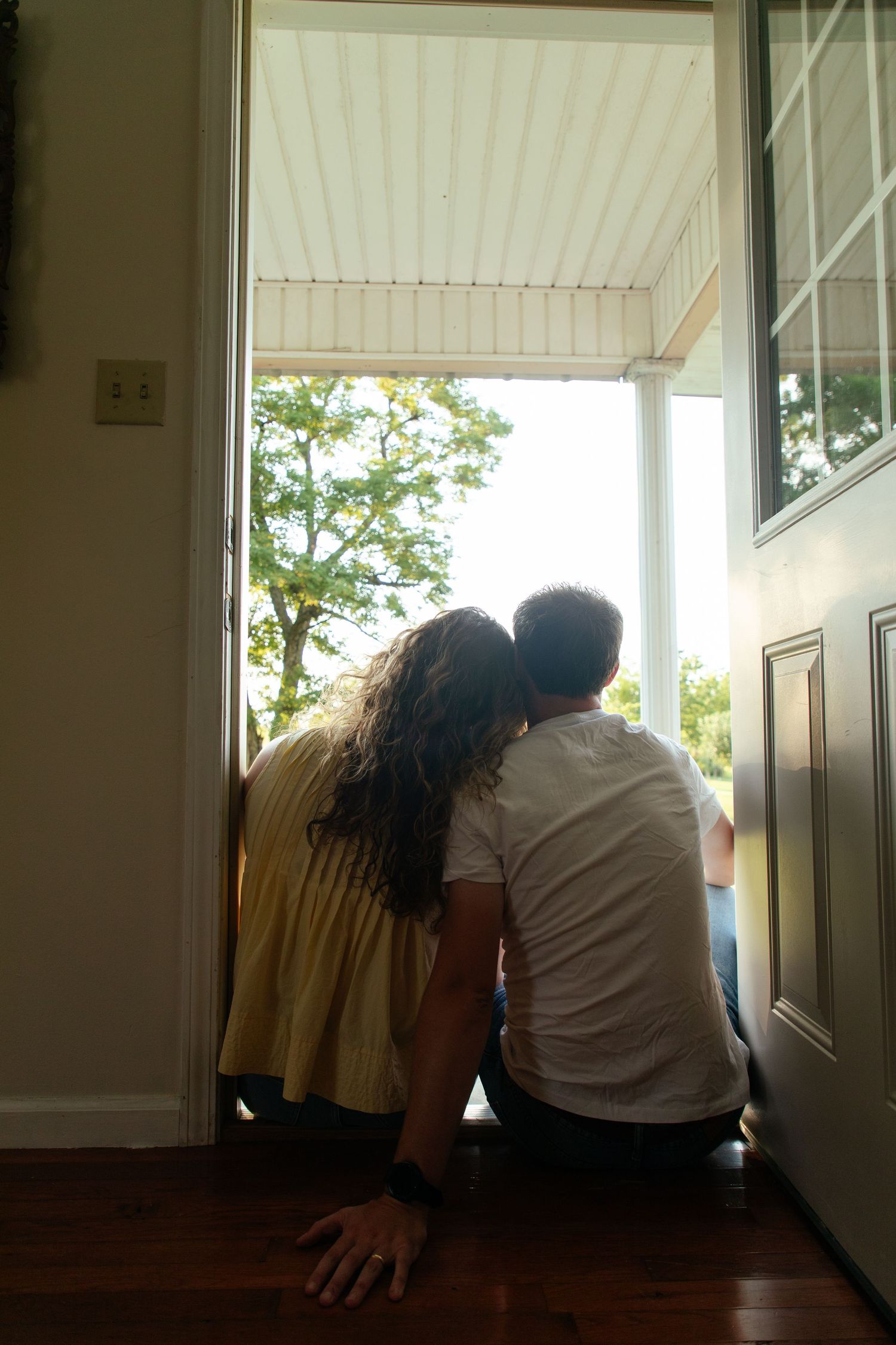 Two people sit close together in silhouette by a window with white trim.