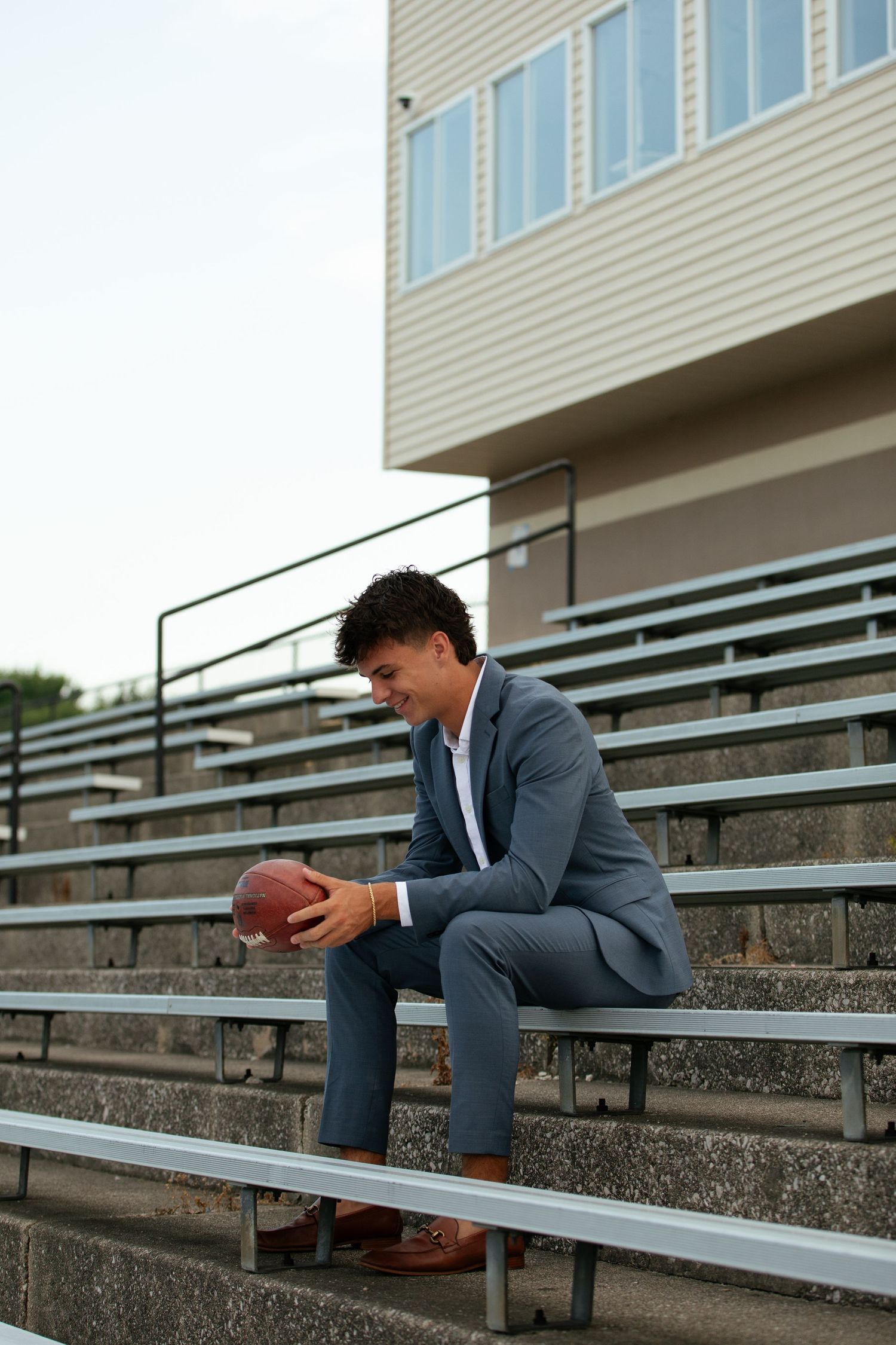 Person in business suit sits alone on outdoor bleachers holding a football.