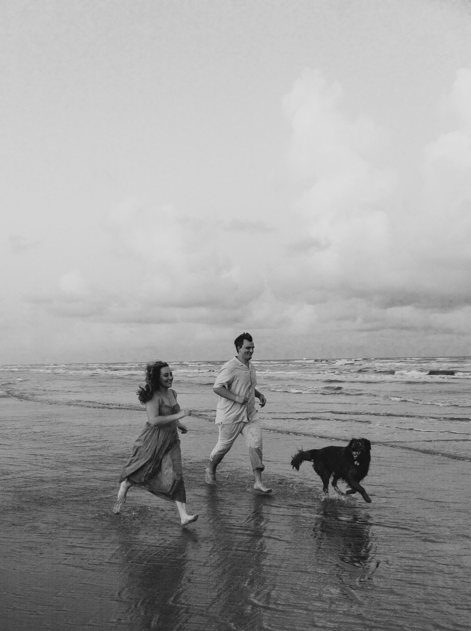 People and dog running together on beach in black and white photo.