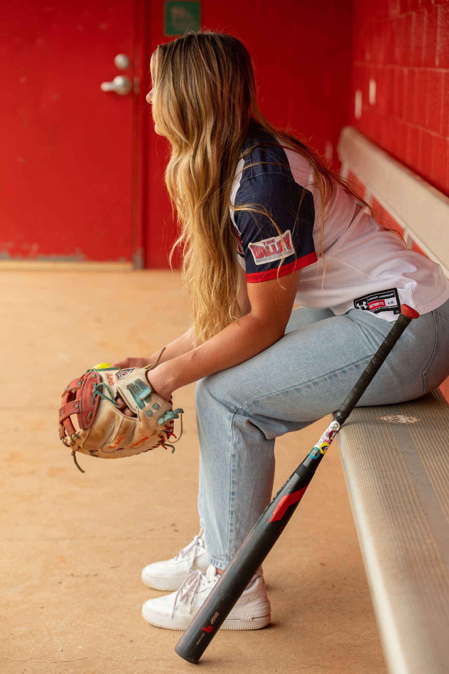 A person in jeans and white sneakers sits on a bench holding a baseball bat.