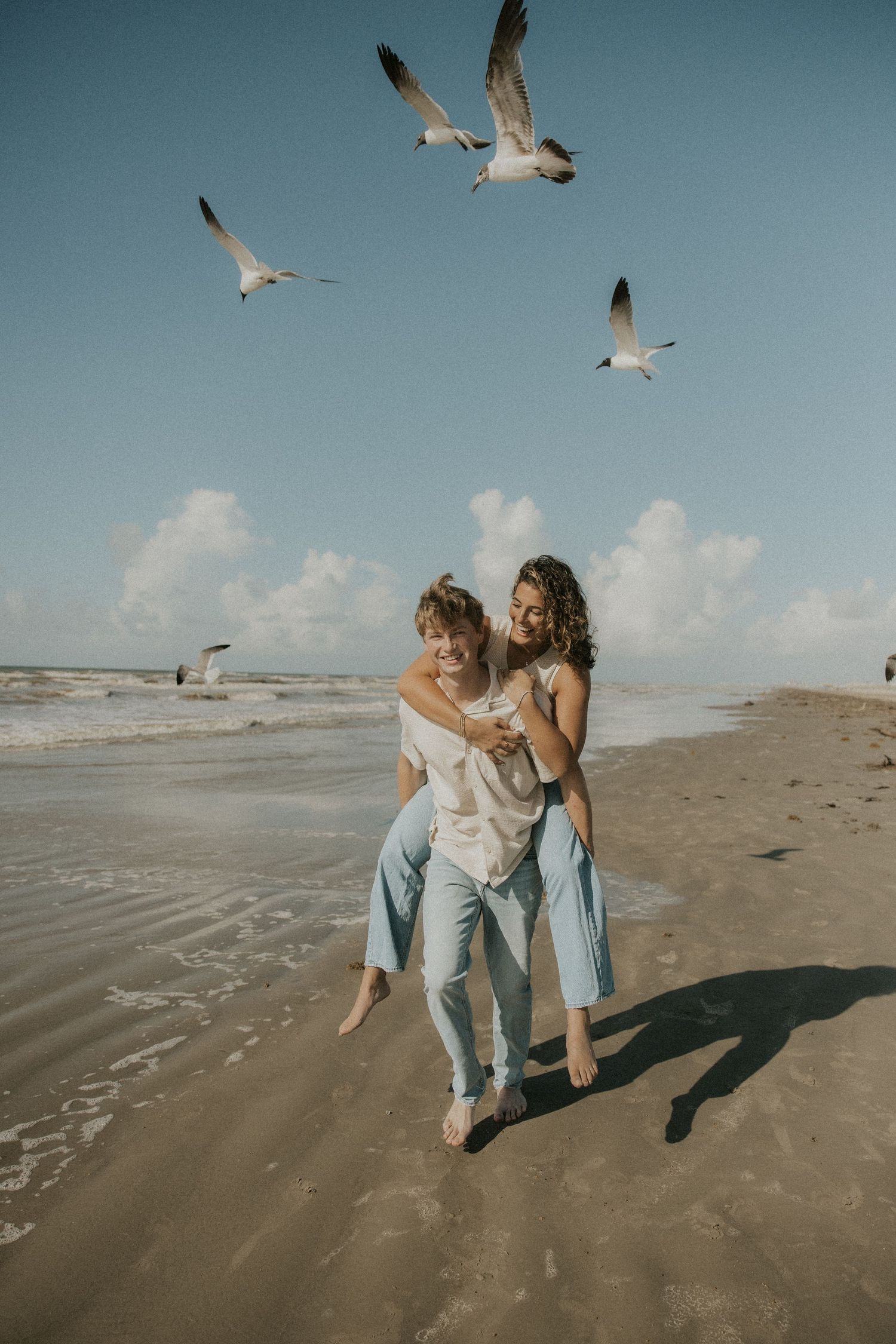 Two people play and dance together on a beach while seagulls fly overhead.