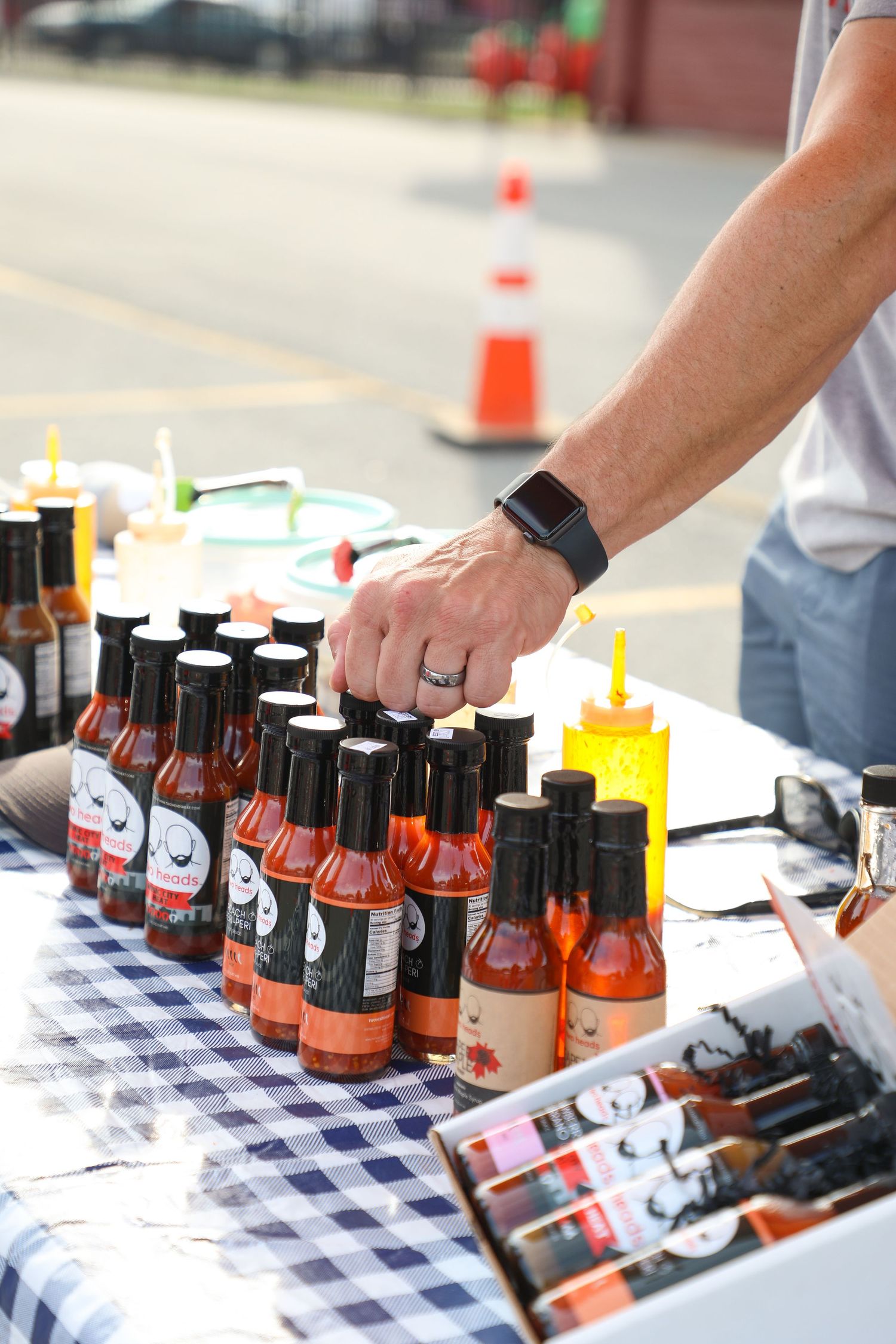 Hot sauce bottles with cartoon face labels arranged on checkered tablecloth at outdoor event.
