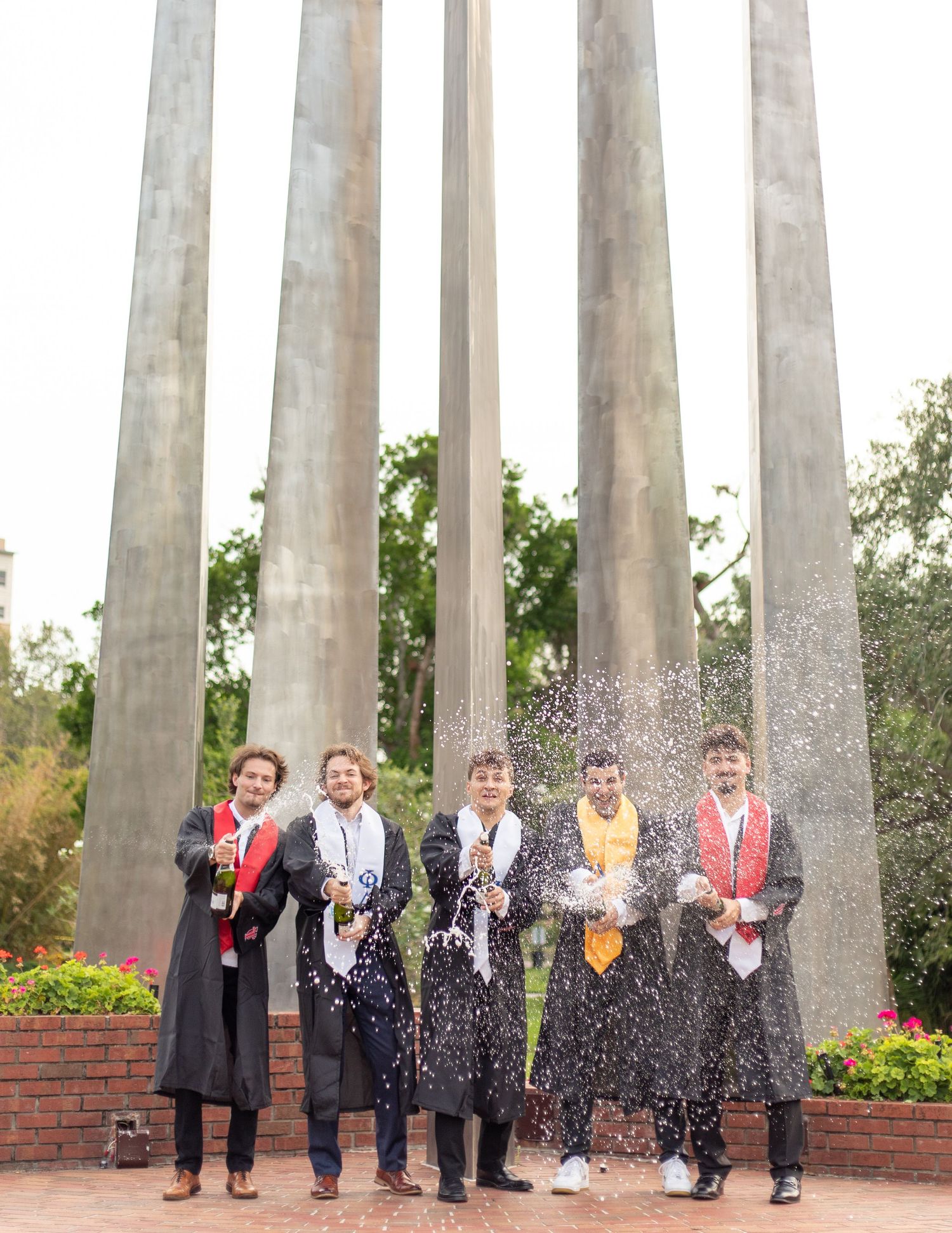 Group celebrates with champagne spray in front of concrete pillars outdoors.