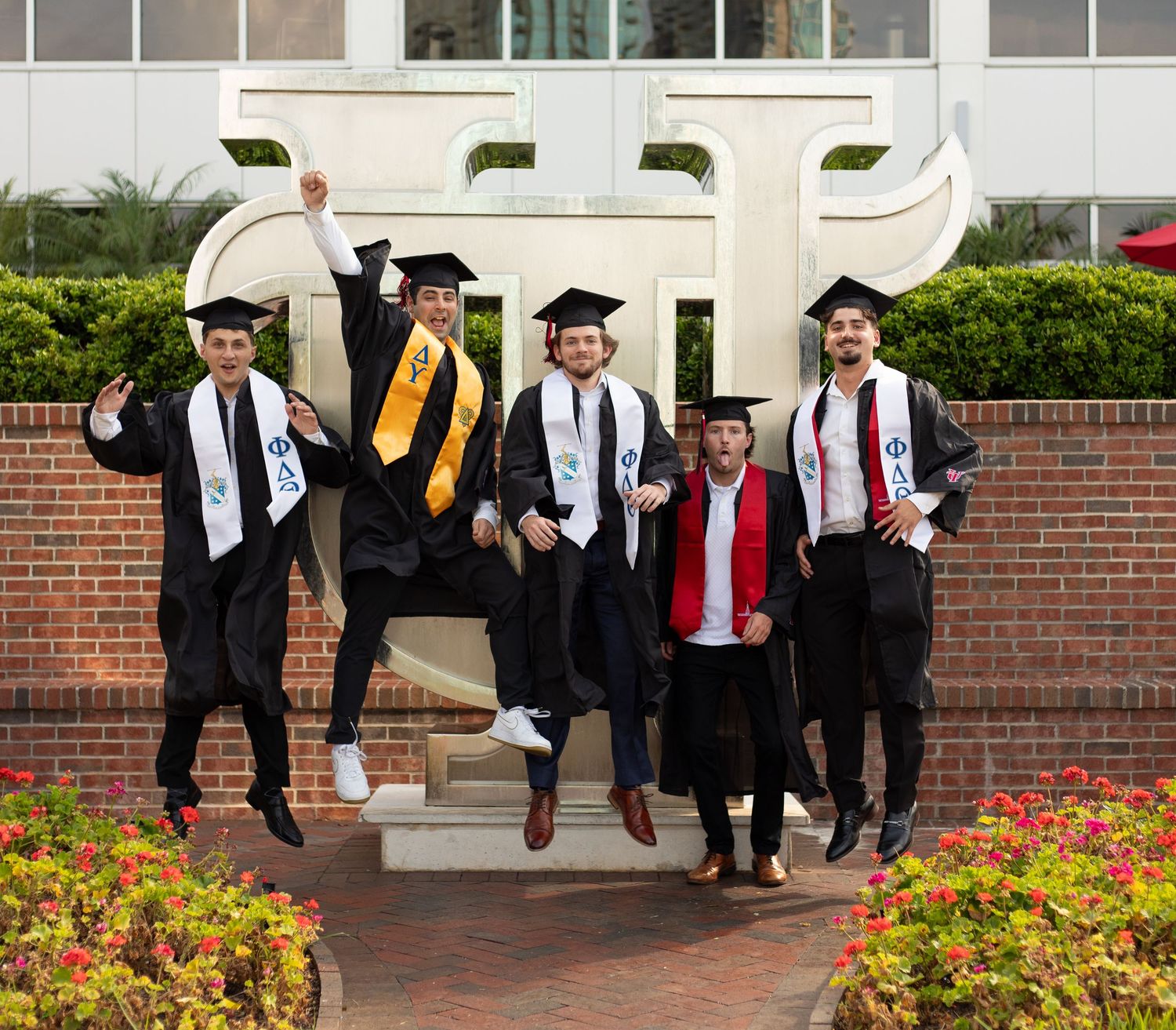 Graduates in black robes and caps jumping with joy outside a brick building.