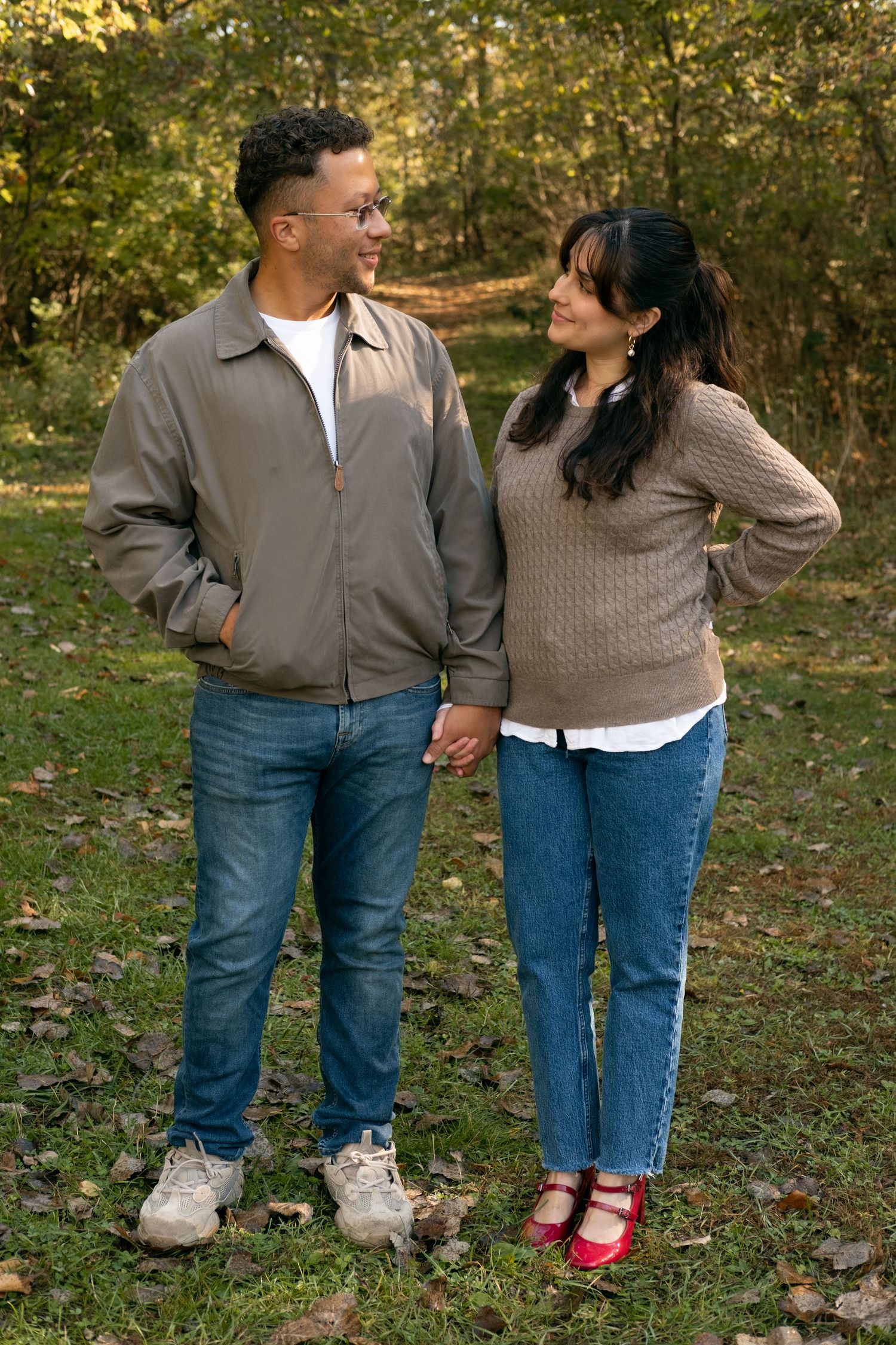 Casual couple in jeans and sweaters pose together among fallen autumn leaves.
