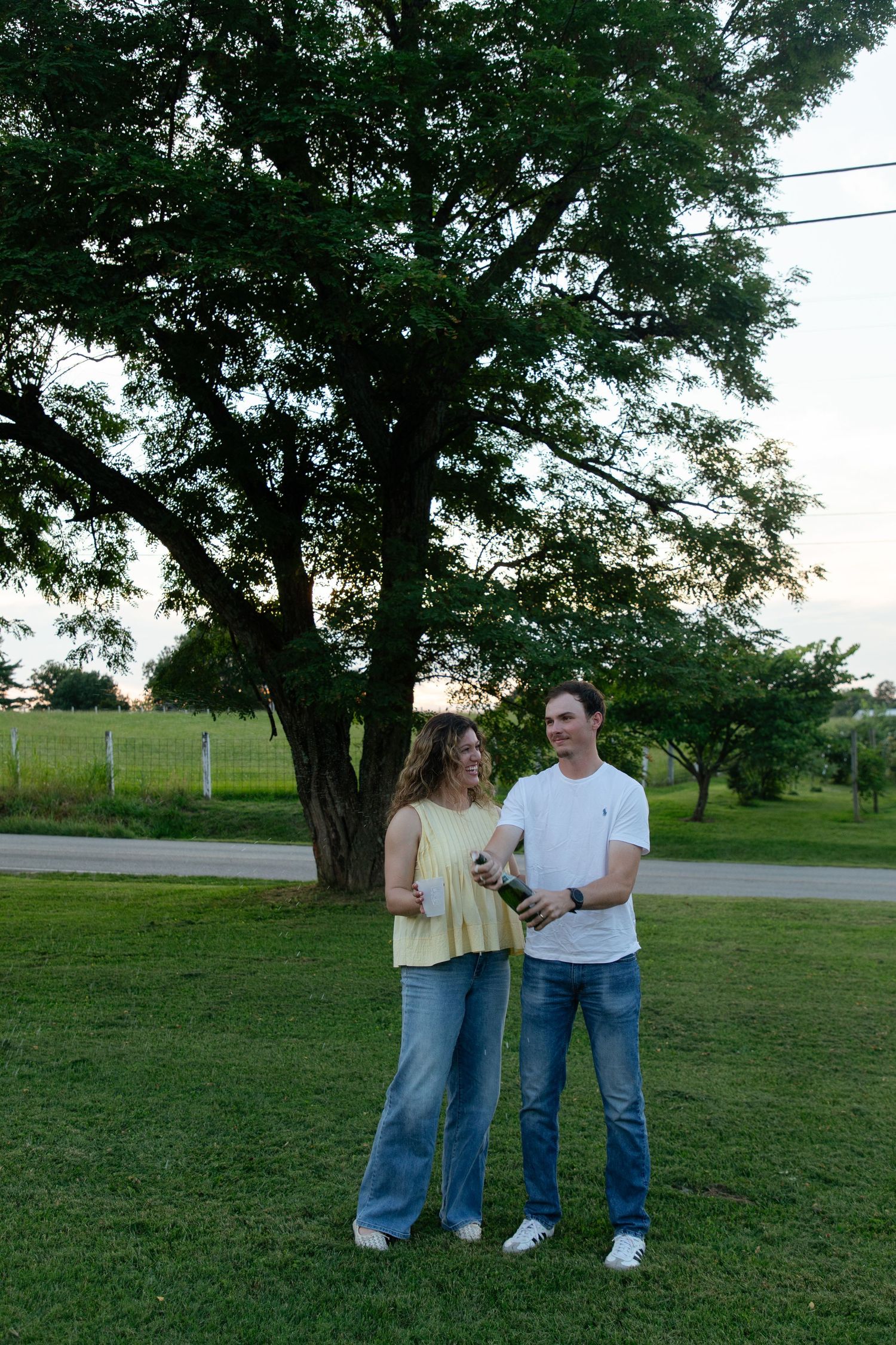 A couple stands together on grass under large trees during golden hour.