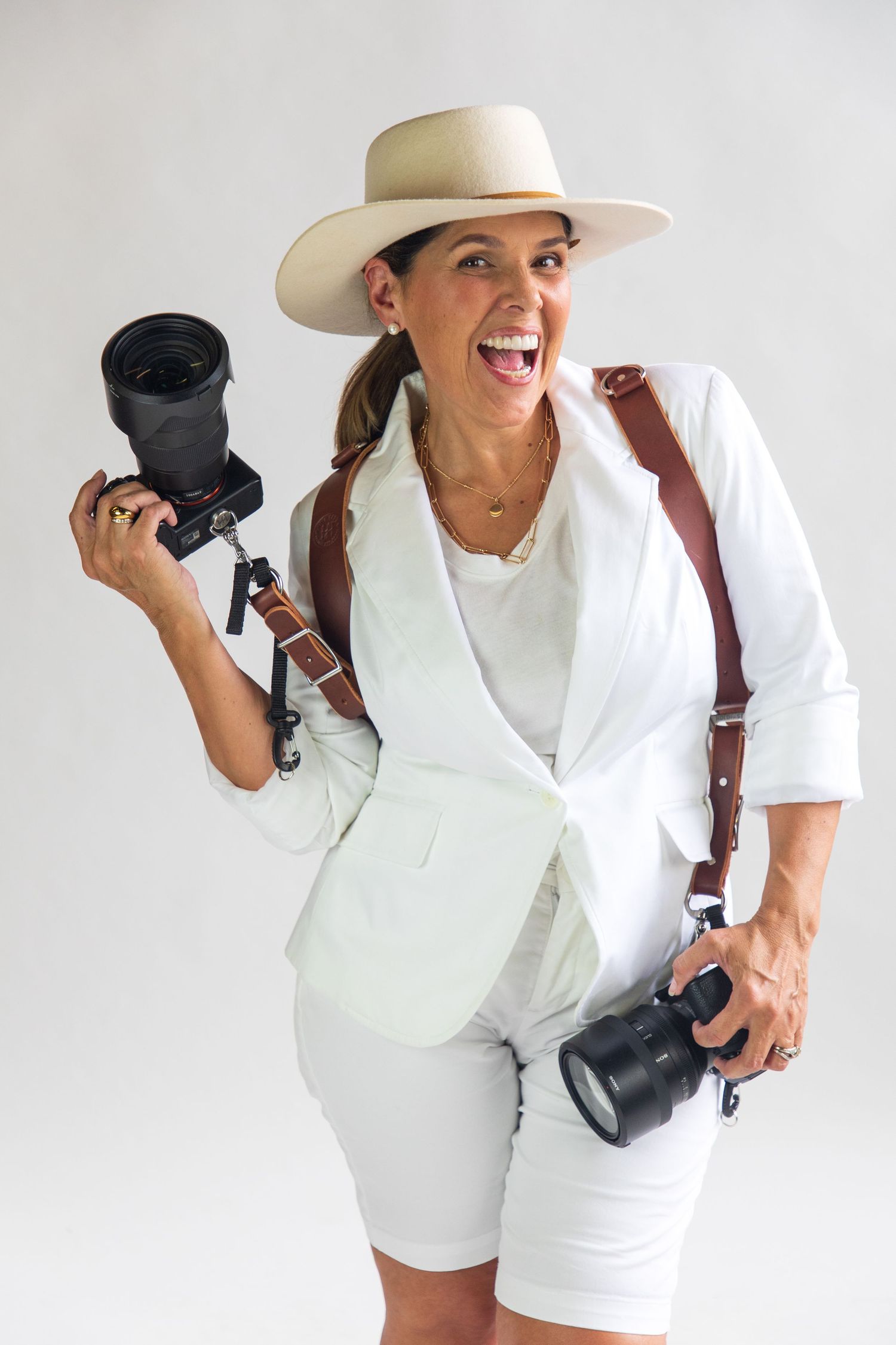 Photographer in white outfit and cowboy hat posing playfully with camera equipment against white backdrop.