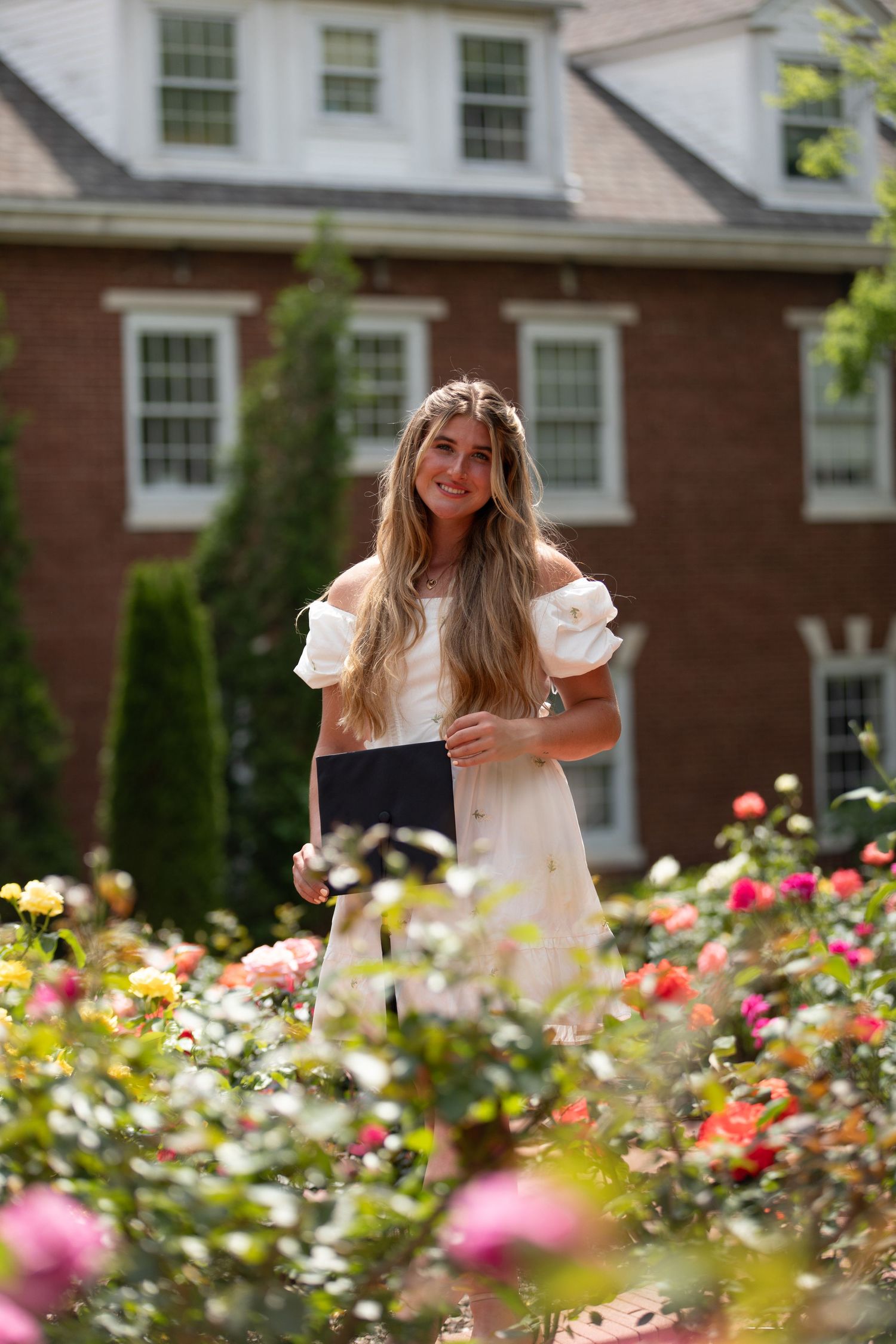 A person poses joyfully in a garden filled with vibrant pink and yellow flowers near a brick building.