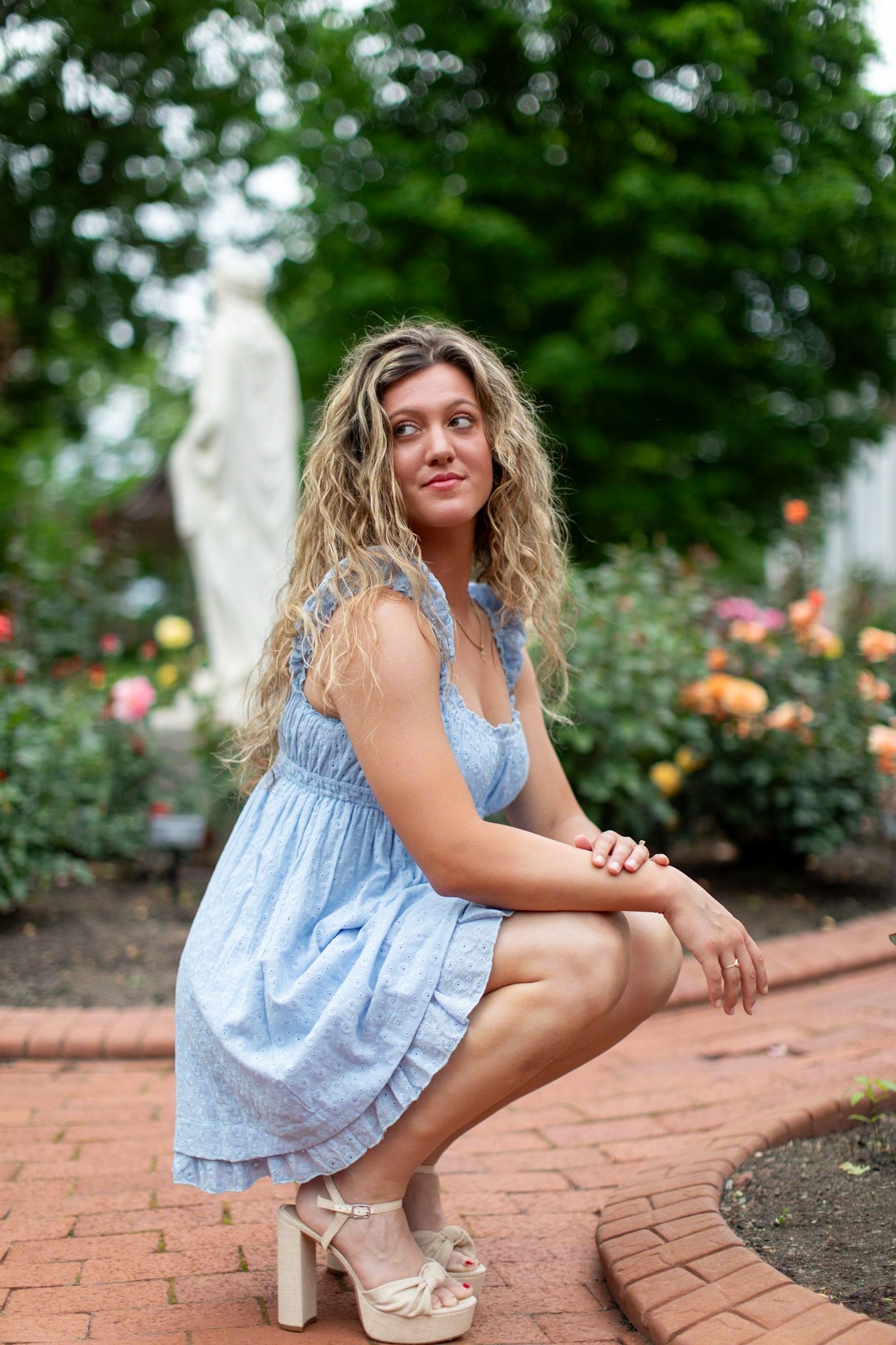 Woman in blue sundress poses on brick pathway near garden.