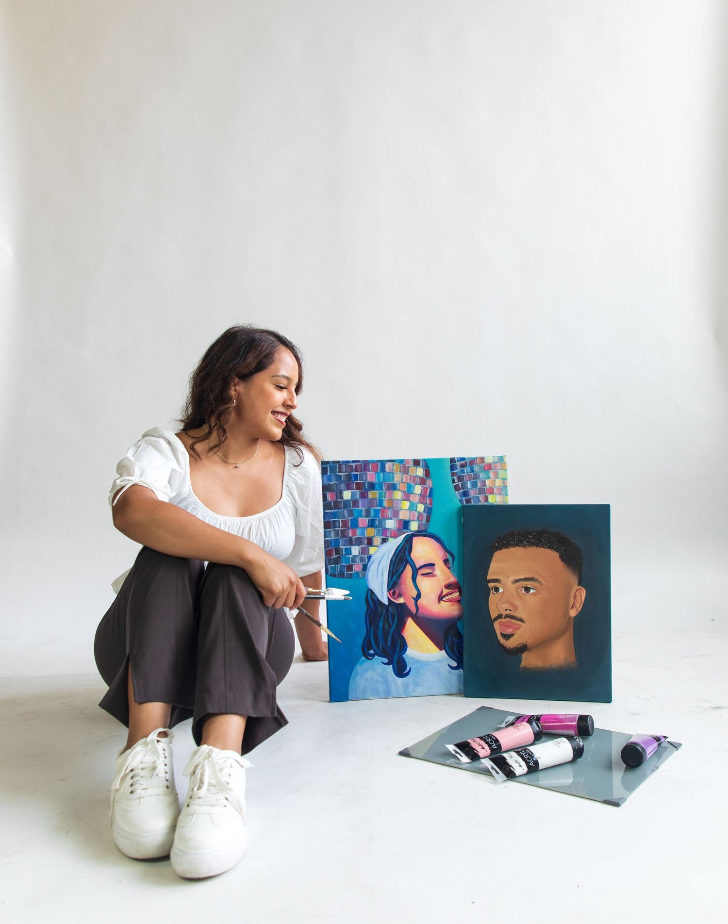 Artist sitting on floor with colorful portrait artwork displays in minimalist white studio setting.