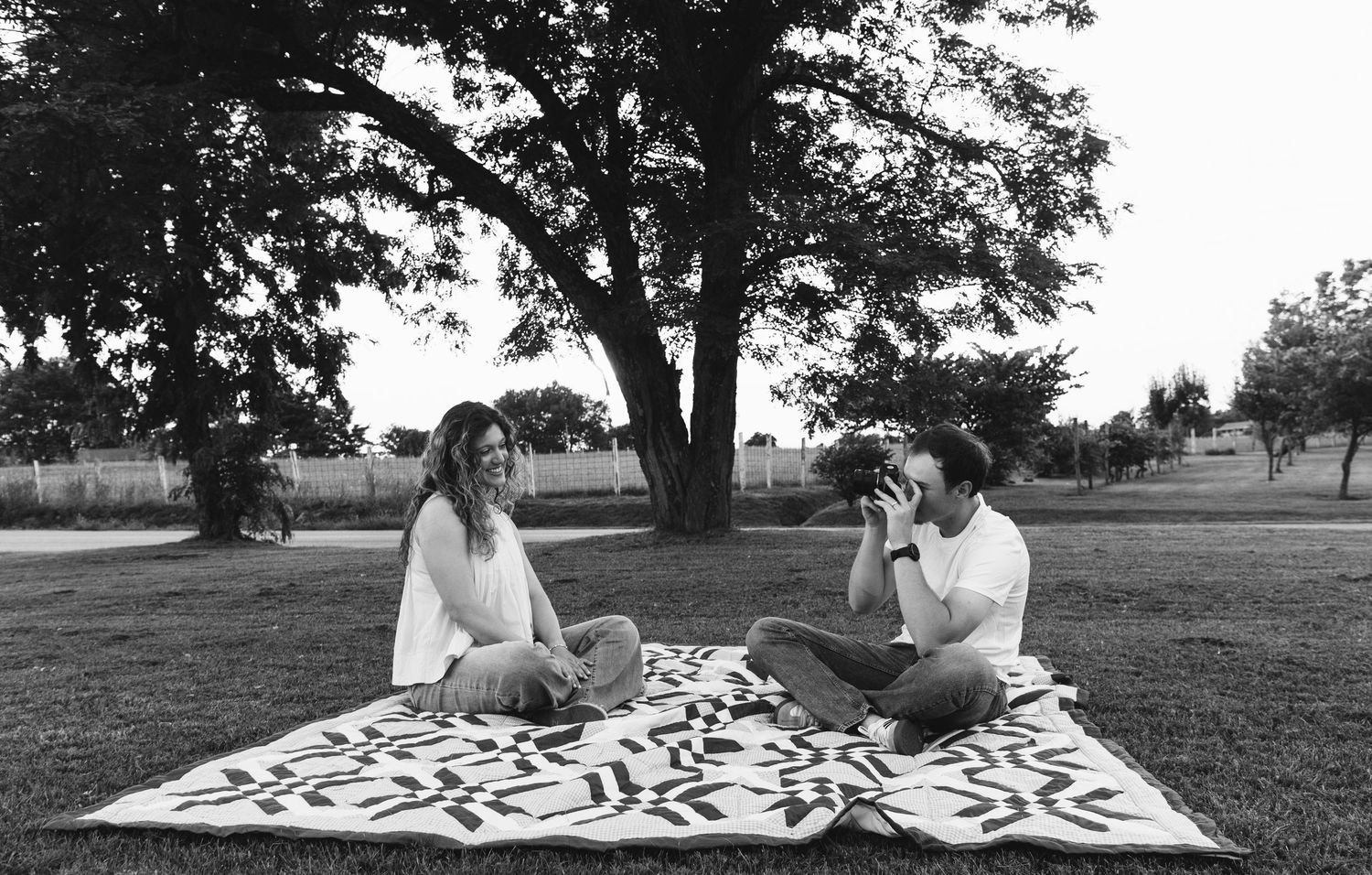 Group relaxing on patterned blanket under large trees in park.
