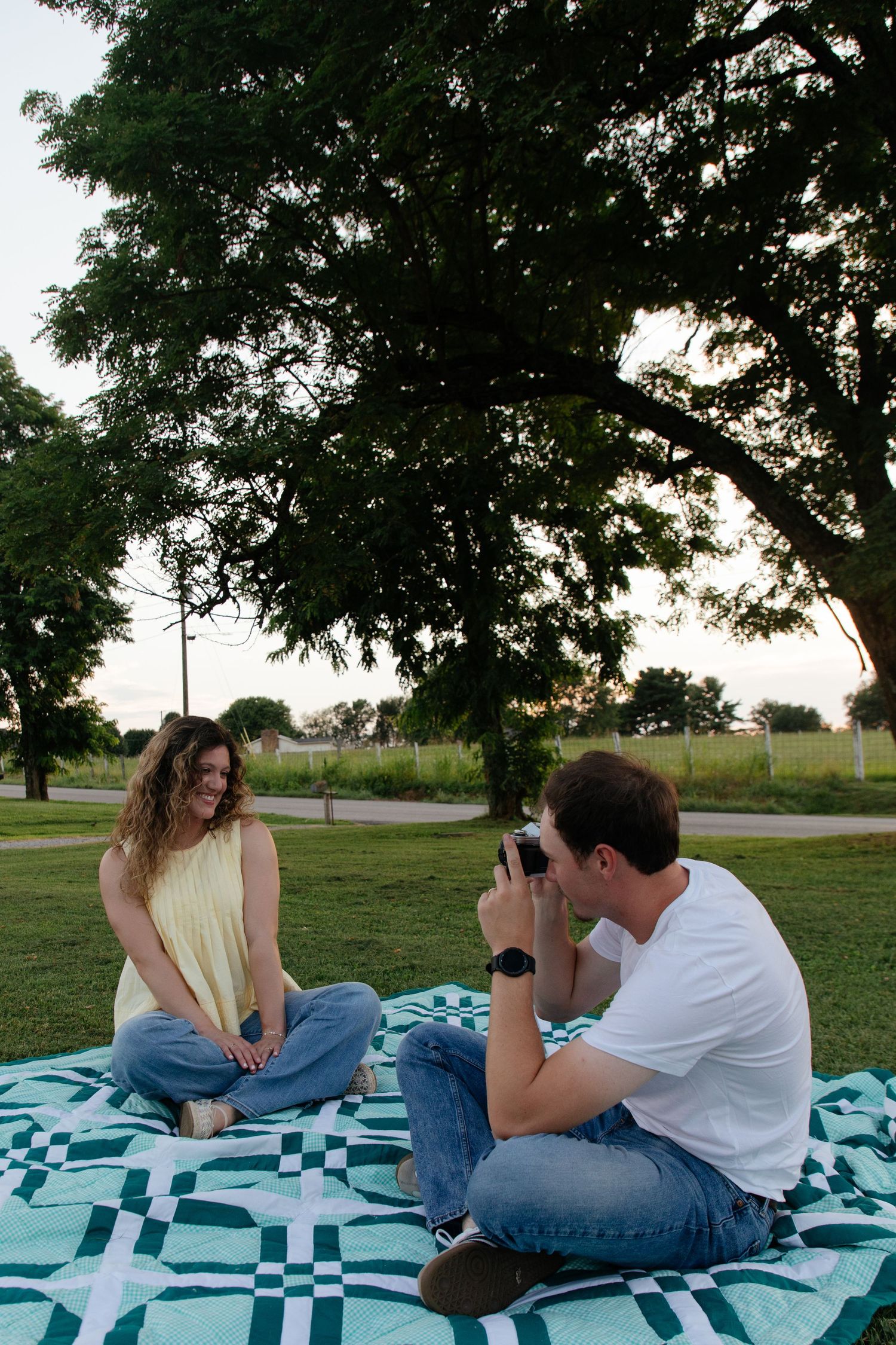 A couple lies on a blue checkered blanket taking photos of each other in a grassy park.