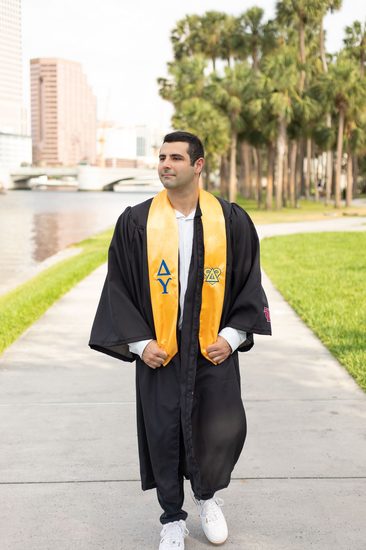 Graduate in black cap and gown with yellow stole walks on campus pathway.