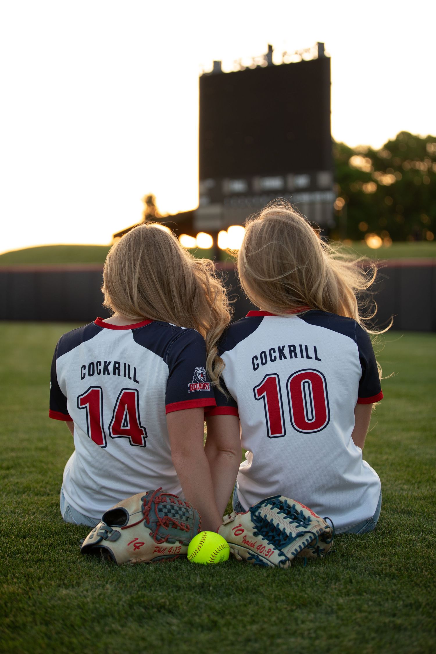 Two softball players in jerseys sit together on field at sunset.