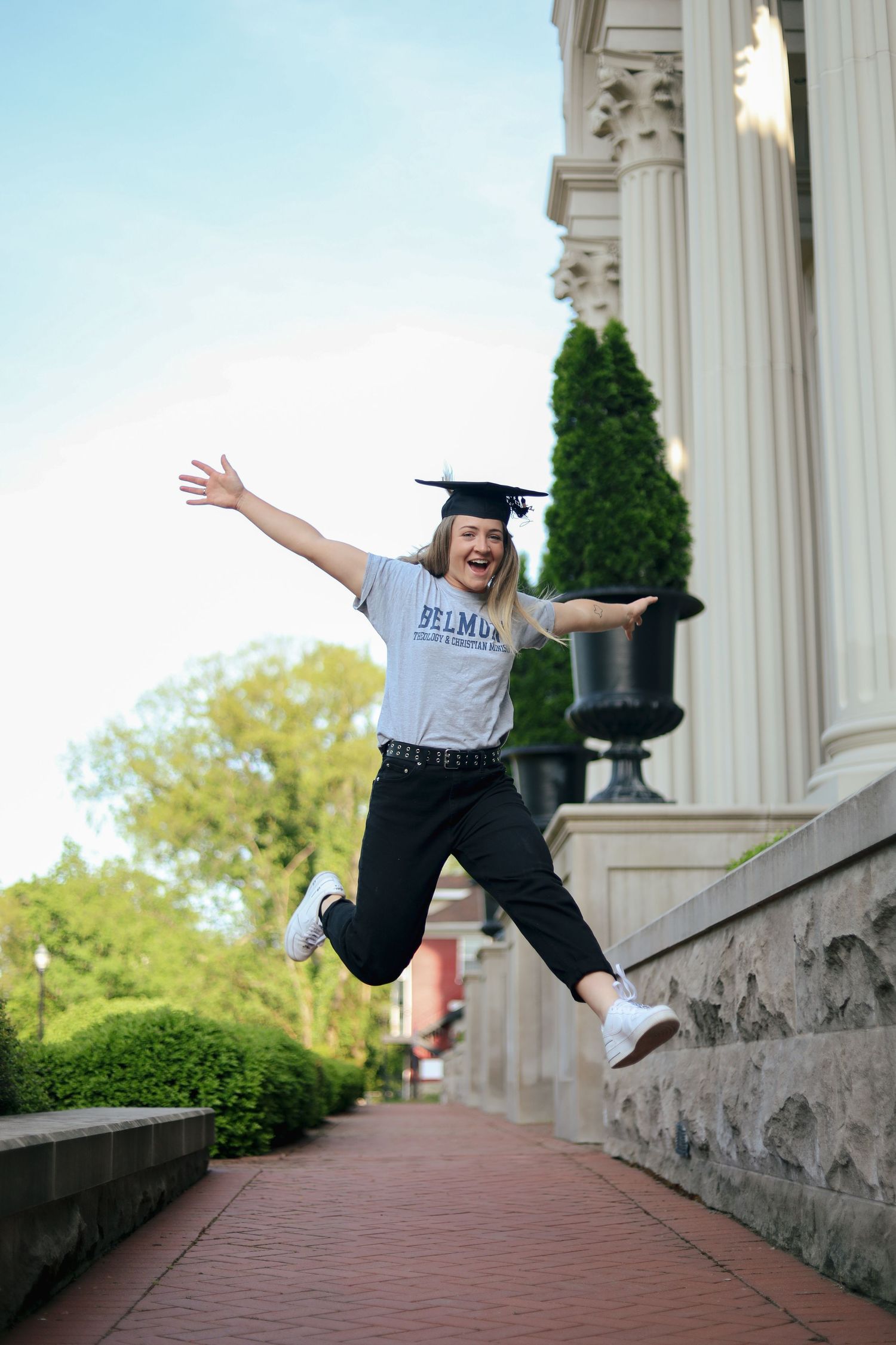 A graduate in cap and gown performs celebratory jumps on a stone walkway with white columns.