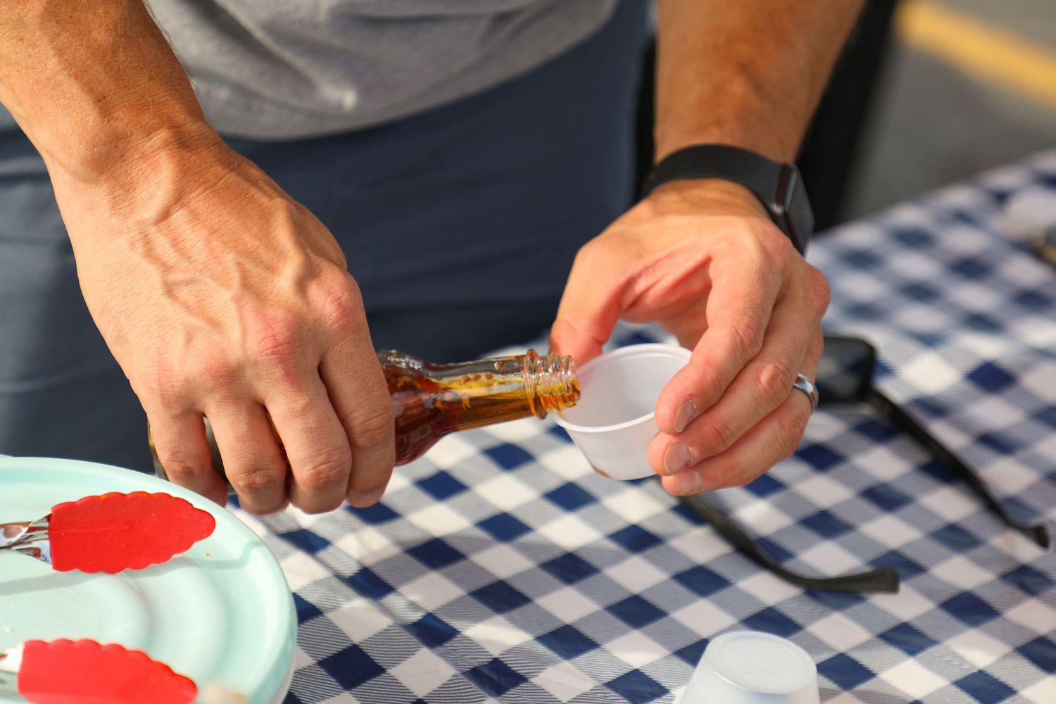 Hands preparing condiment samples on blue checkered tablecloth at outdoor market display.