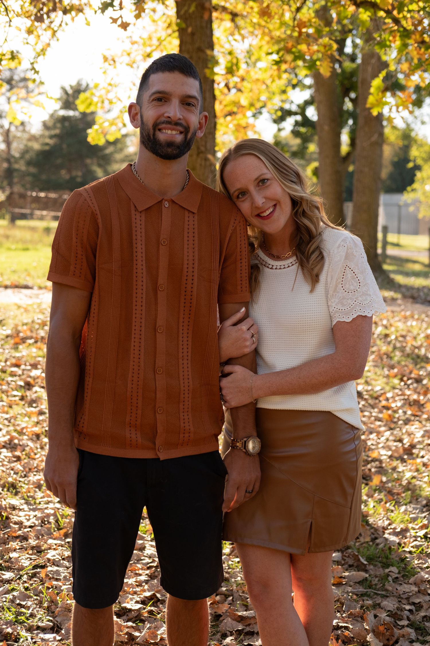Couple in autumn outfits laughs together during sunset photoshoot with fallen leaves.