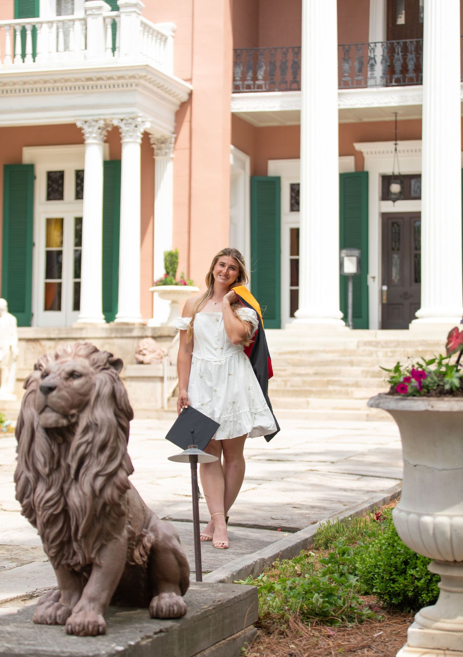 Woman in white dress poses near stone lion statue and pink building.