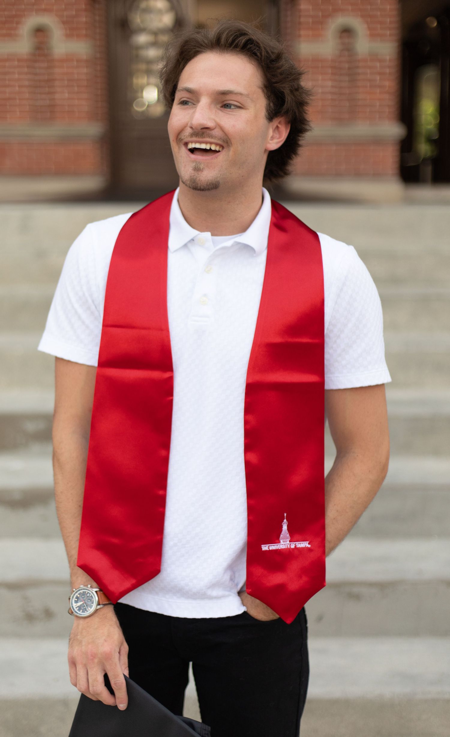 Graduate wearing red stole poses casually in front of brick university building.