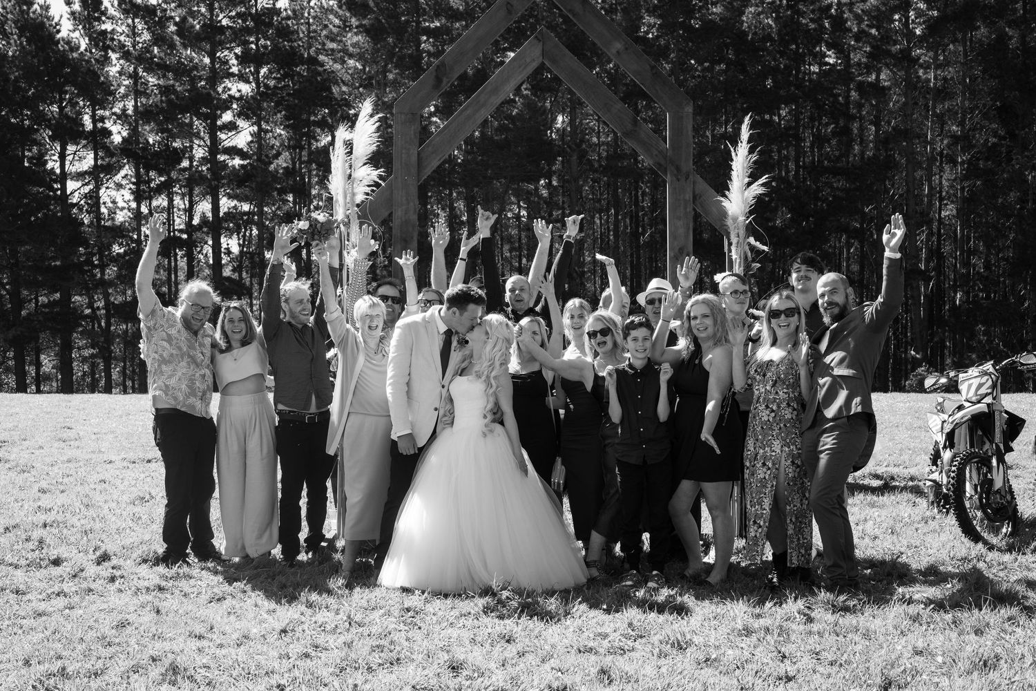 Bride and groom kissing under a wooden arch while surrounded by cheering wedding guests with hands raised in celebration.