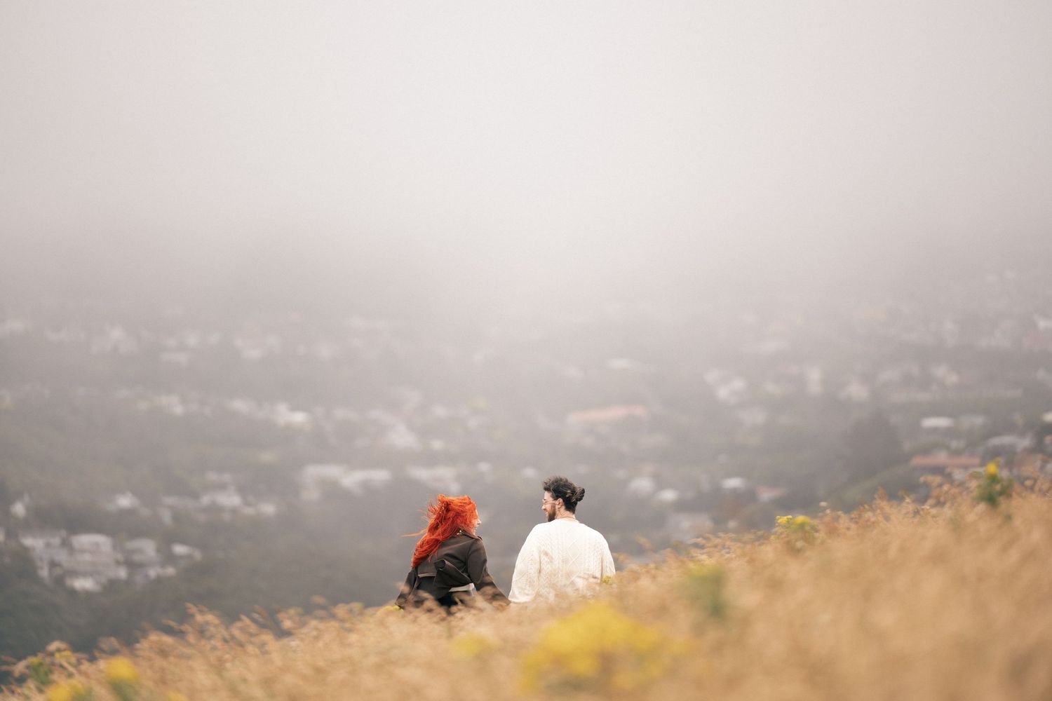Engaged couple photoshoot on golden grassy hillside with a misty city landscape in the background.