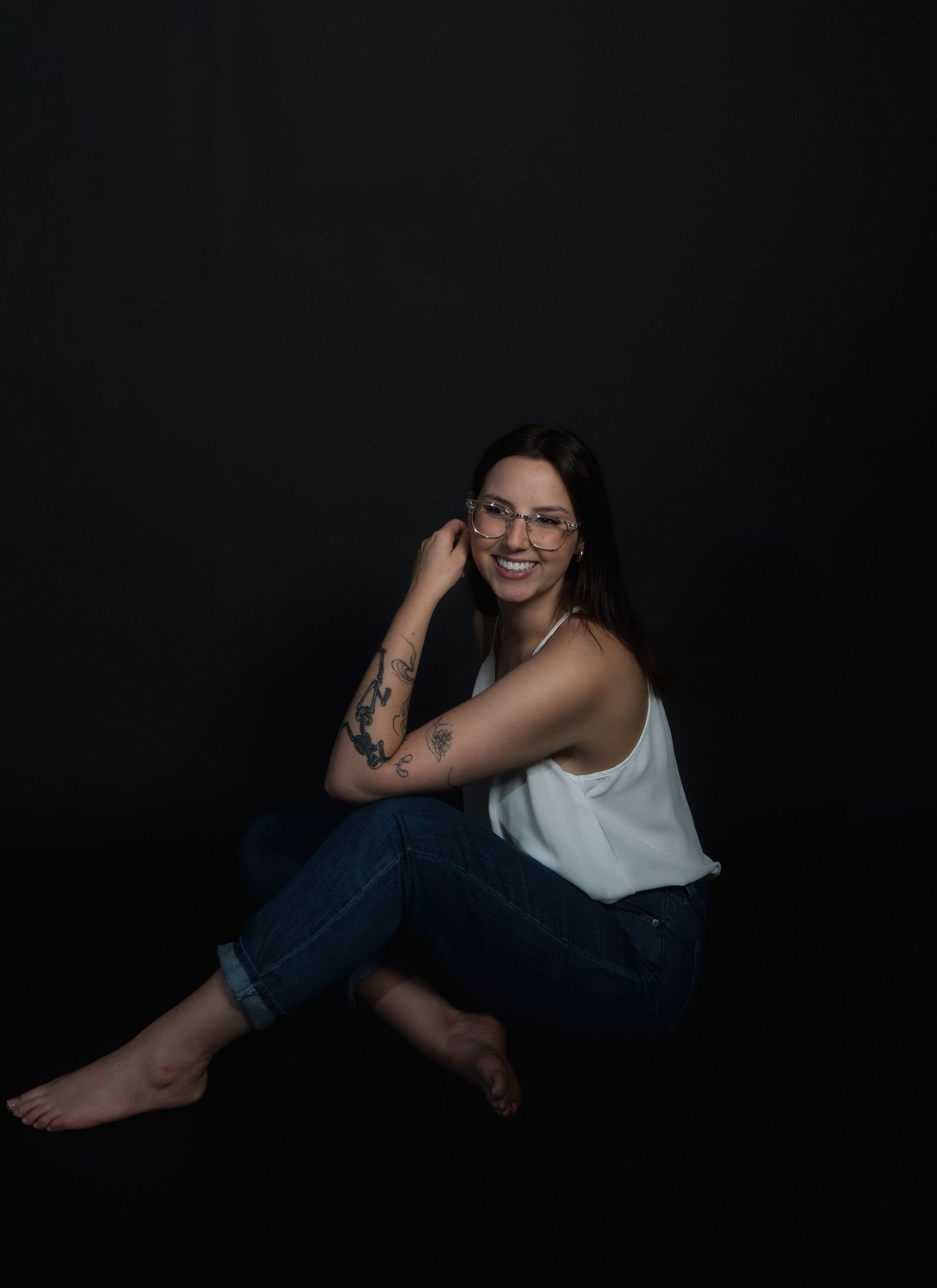 Smiling brunette woman with glasses and tattoos sitting on floor against black backdrop.