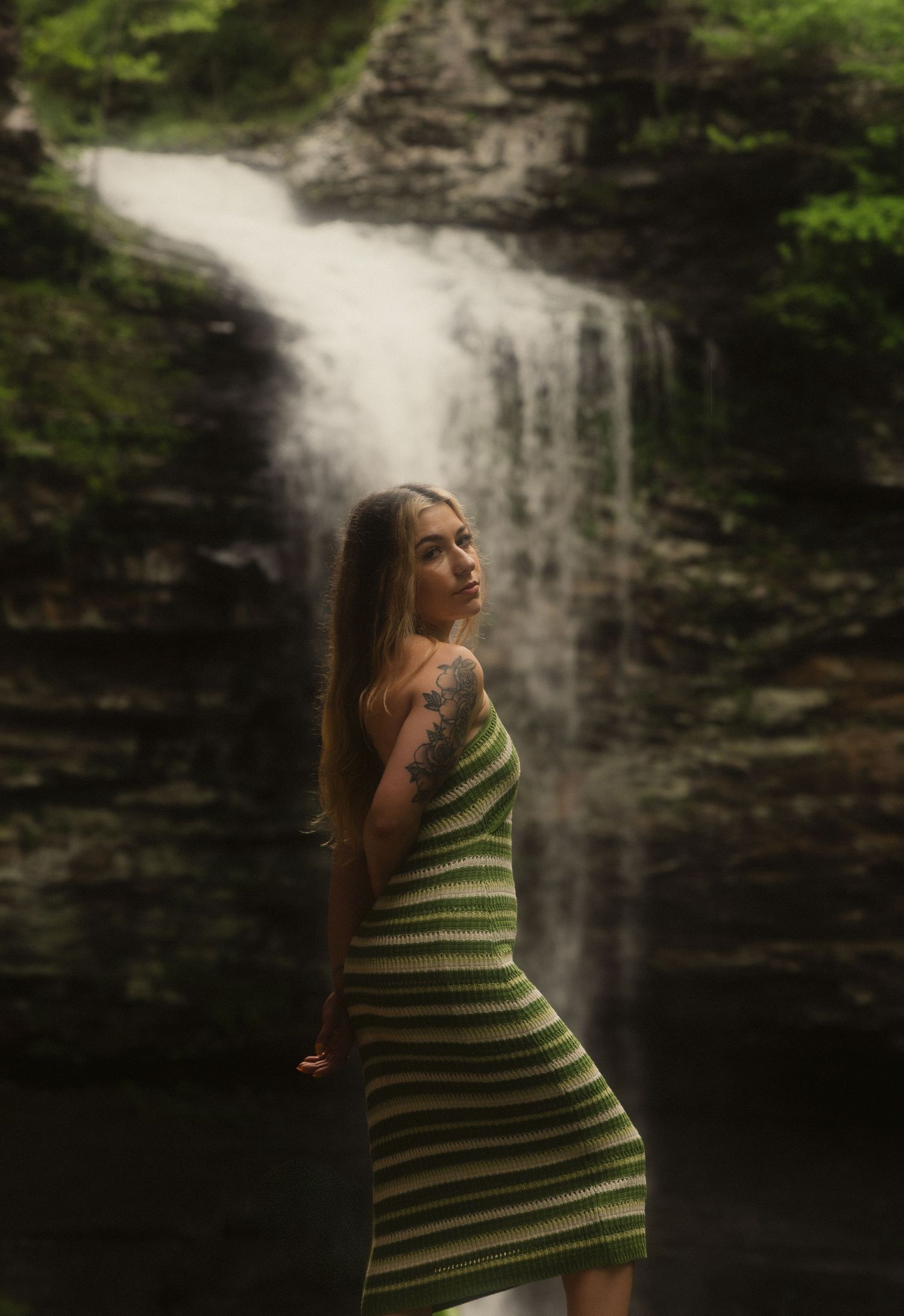 A woman in a green striped dress stands before a cascading waterfall surrounded by lush greenery.