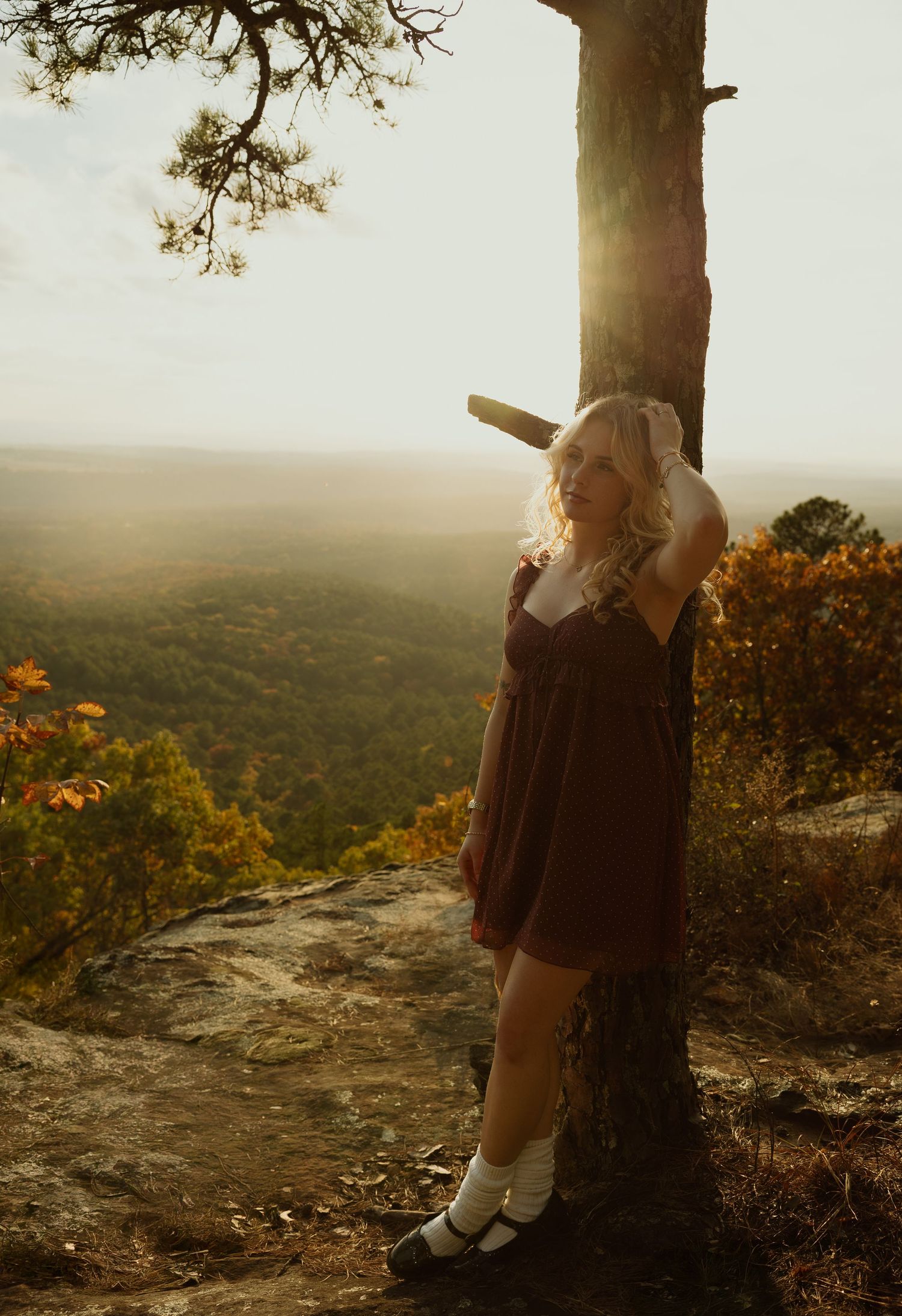 A blonde woman in a burgundy dress leans against a pine tree at sunset on a scenic rocky overlook.