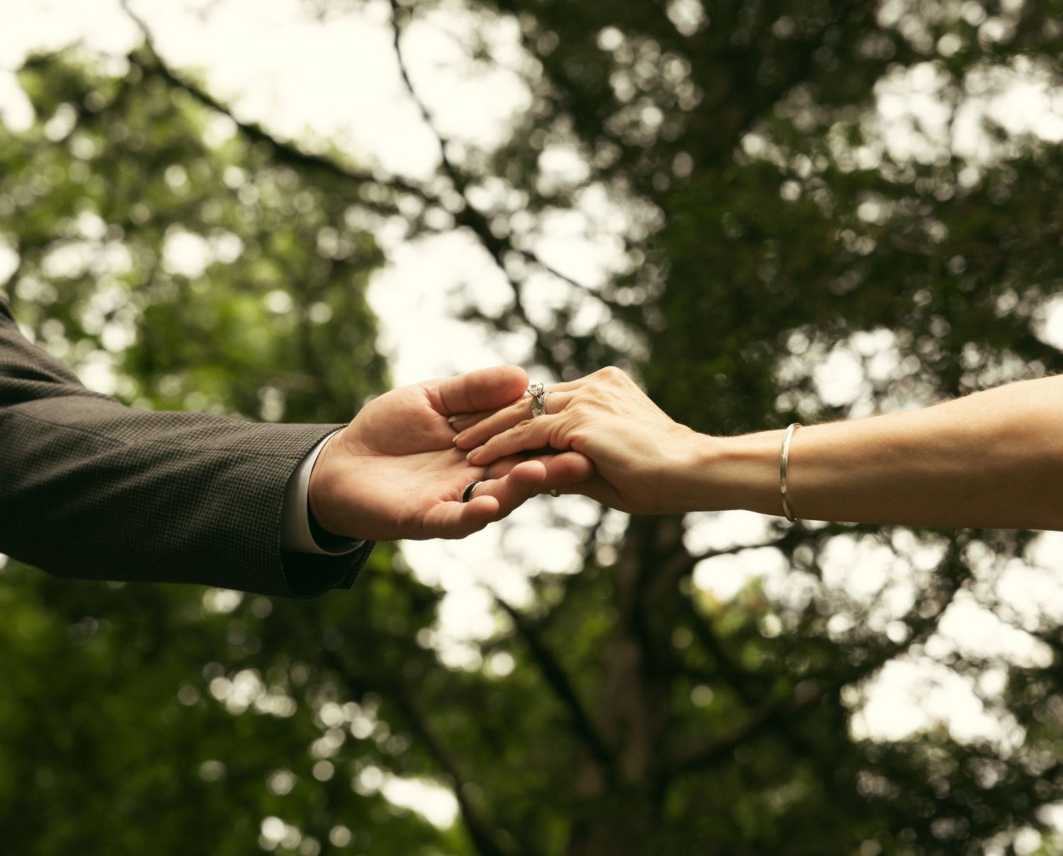 Close-up of a groom's hand holding a bride's hand with a ring on her finger, set against softly blurred green trees.