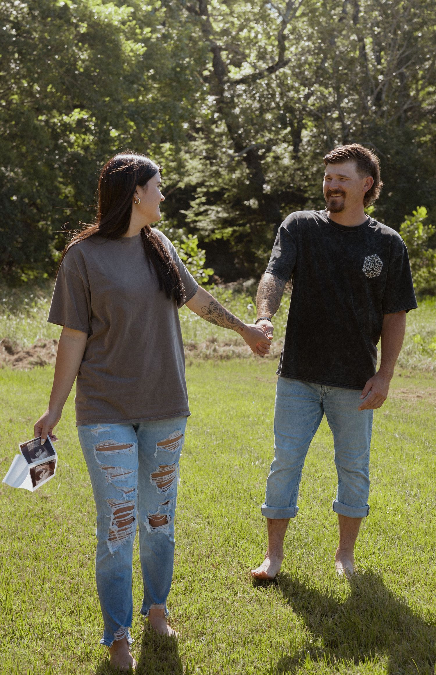 Smiling couple holding hands and walking in sunny green yard while woman holds ultrasound photos.