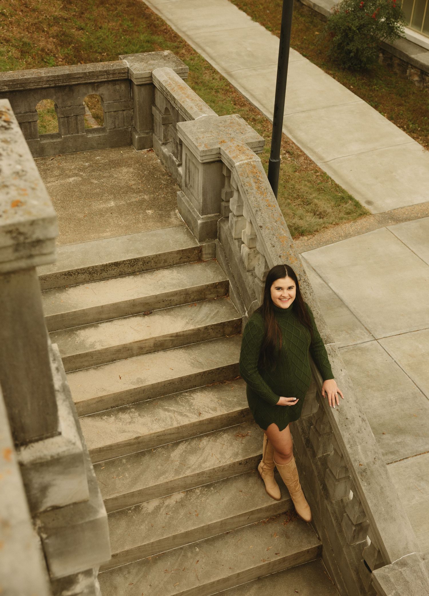 Pregnant woman in green dress and tan boots posing on stone steps outdoors.