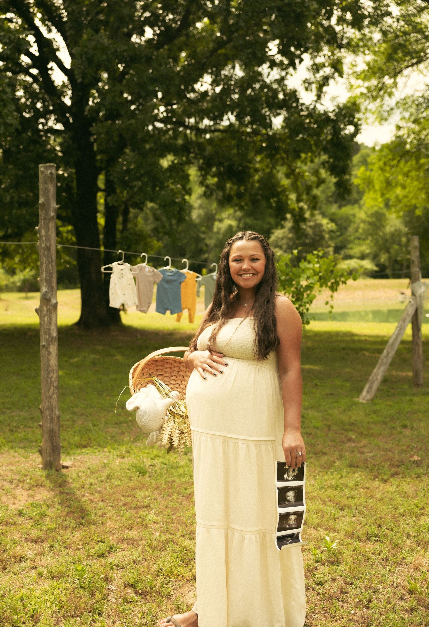 Pregnant woman in yellow maxi dress holding ultrasound photos and basket outdoors with baby clothes on clothesline.