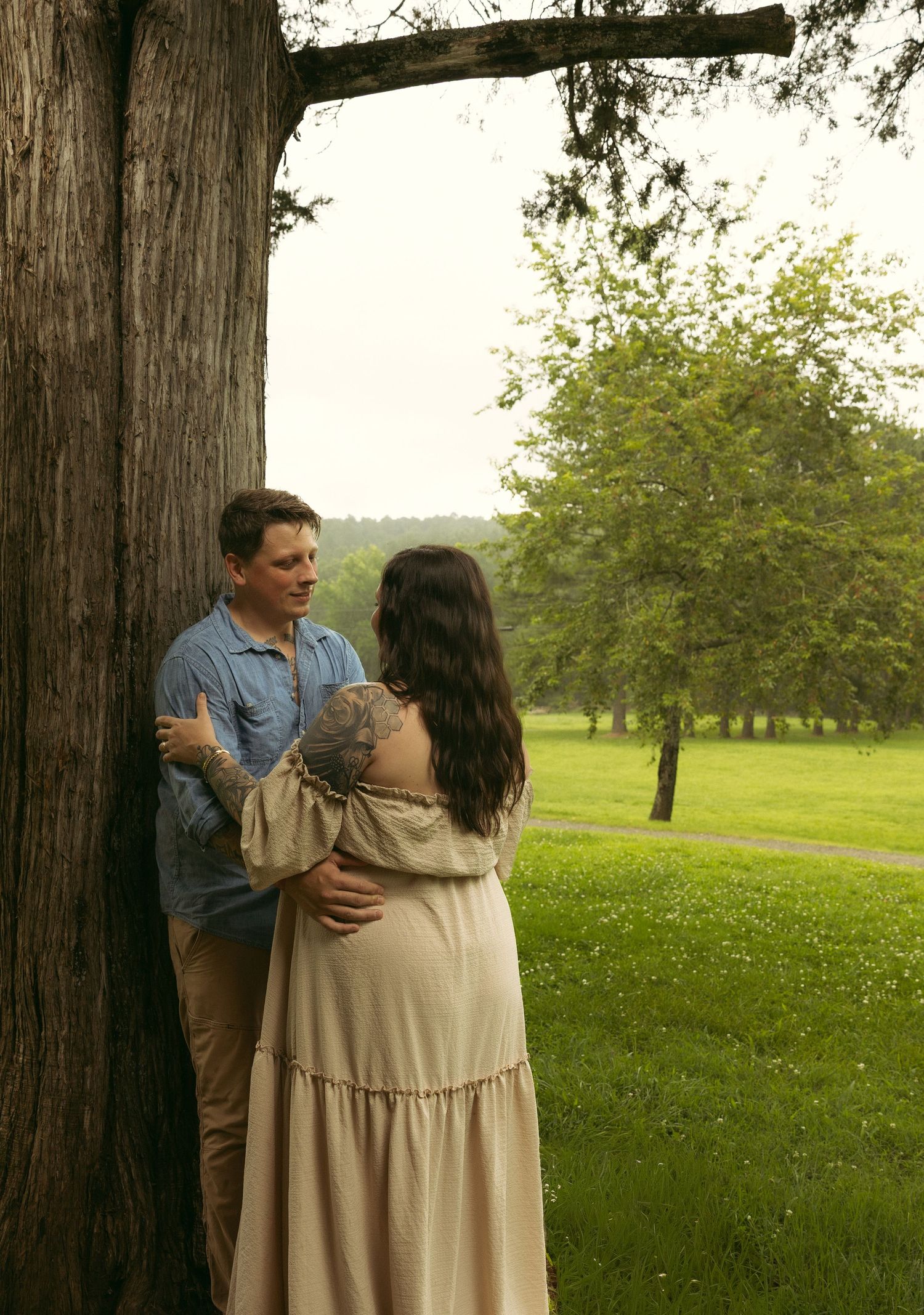 A couple shares a romantic moment by a large tree in a lush green park, woman in beige dress, man in blue shirt.